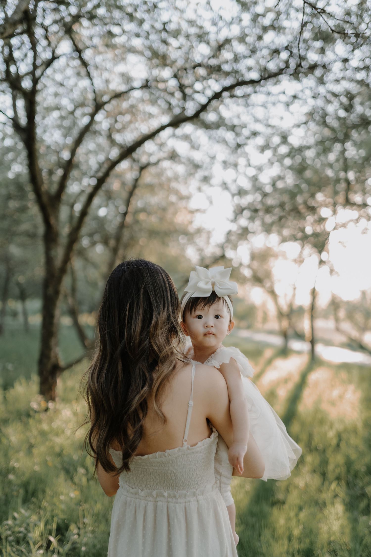 A woman in a white dress holds a baby in a sunny outdoor setting with trees at sunset.