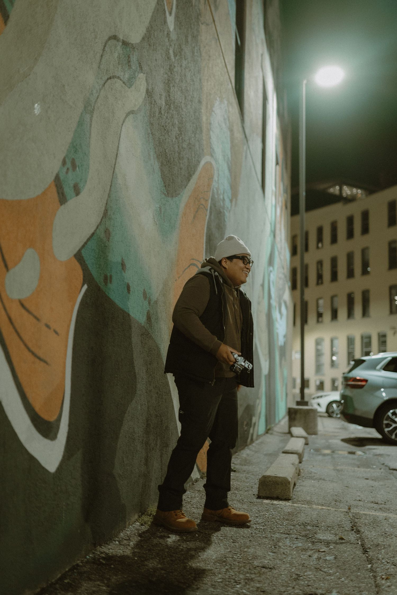 Photographer in beanie and sunglasses holding camera at night beside colorful street mural in urban alley.