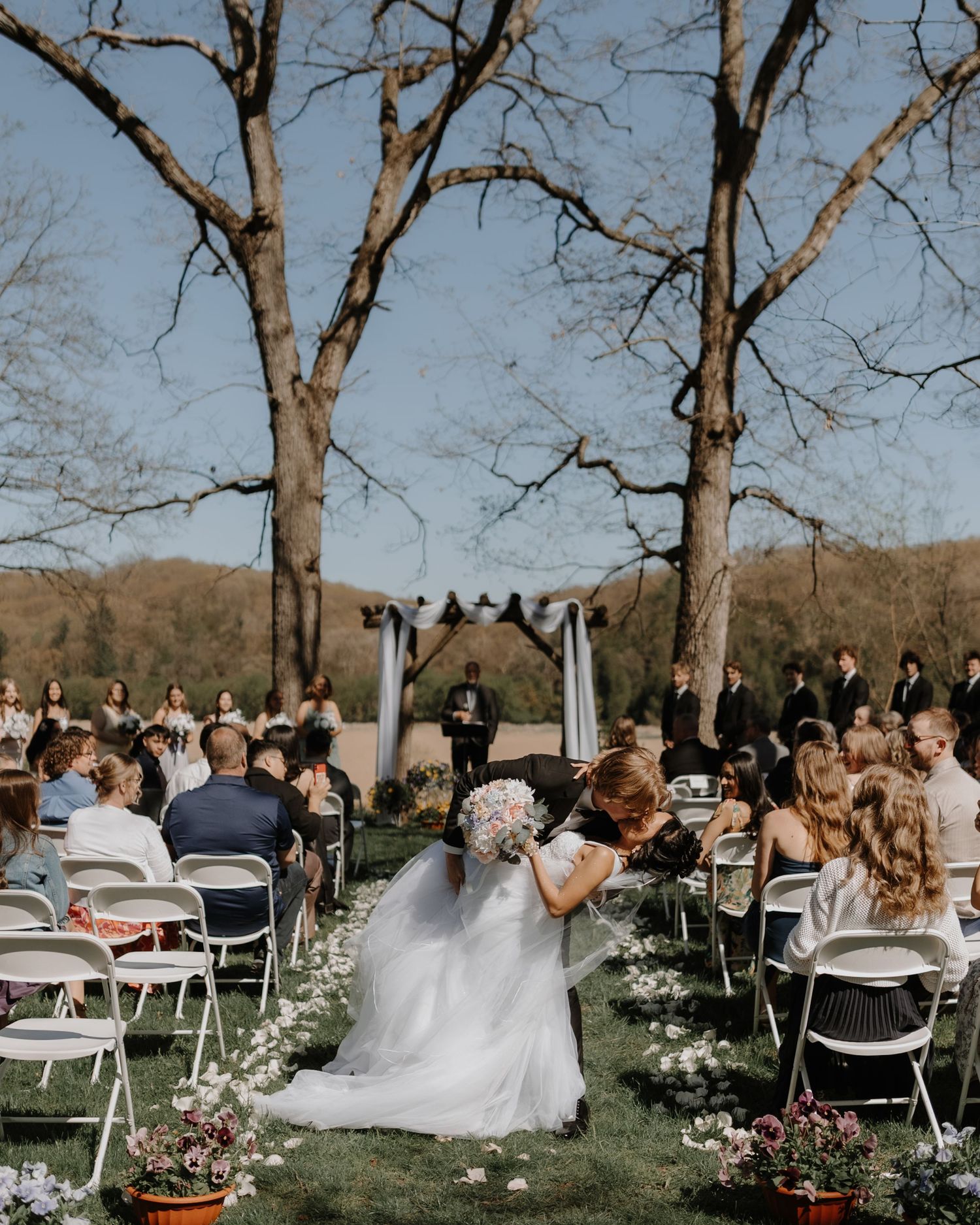 Guests watch a celebration at an outdoor wedding venue with white chairs and floral arrangements.