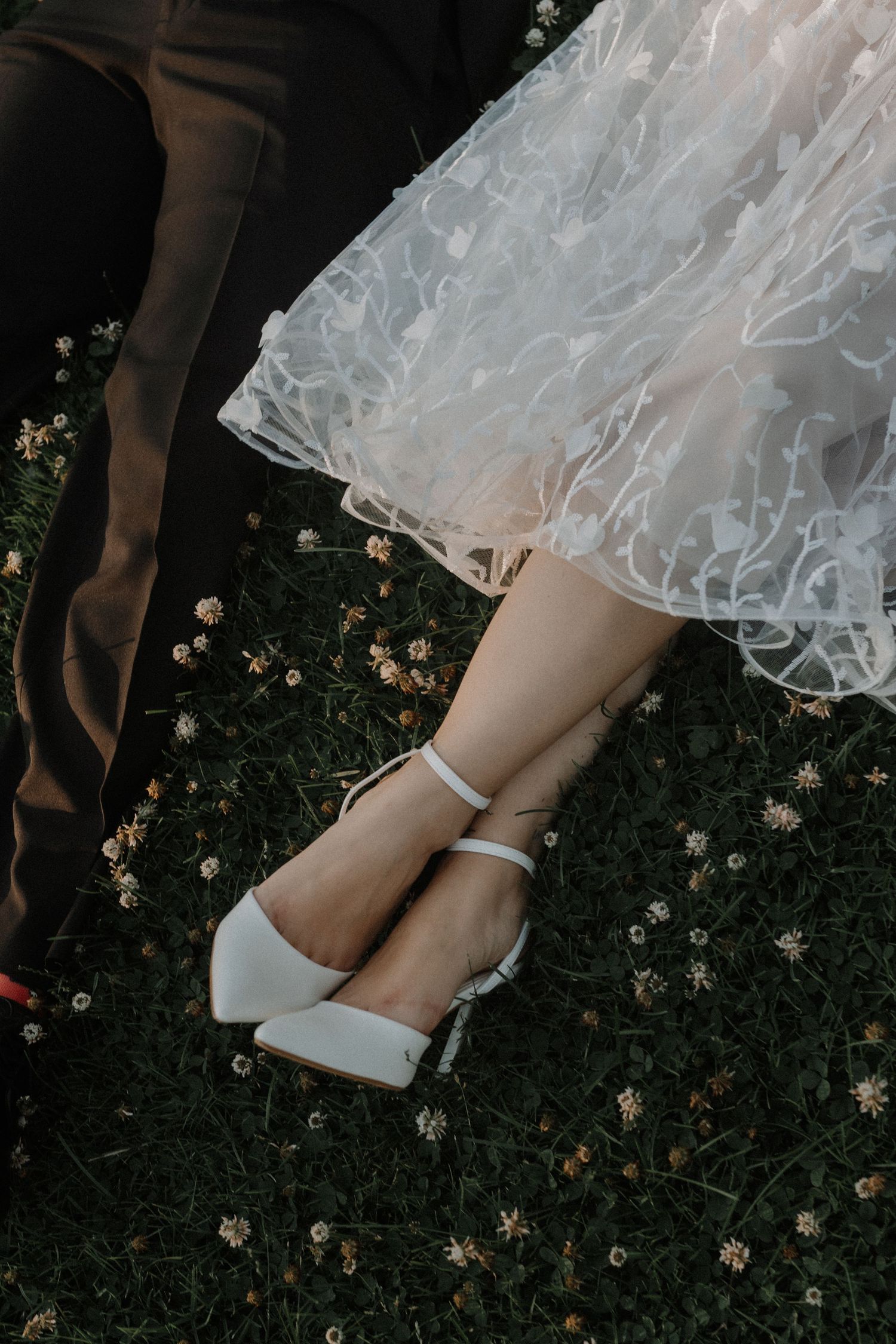 White tulle dress and elegant ankle-strap shoes against dark grass with scattered small flowers.