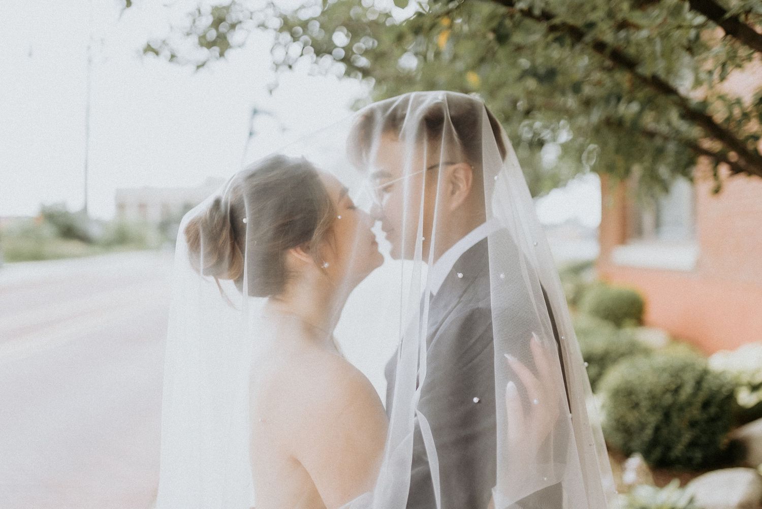 A romantic wedding couple stands close together under a delicate white veil on a sunny day.