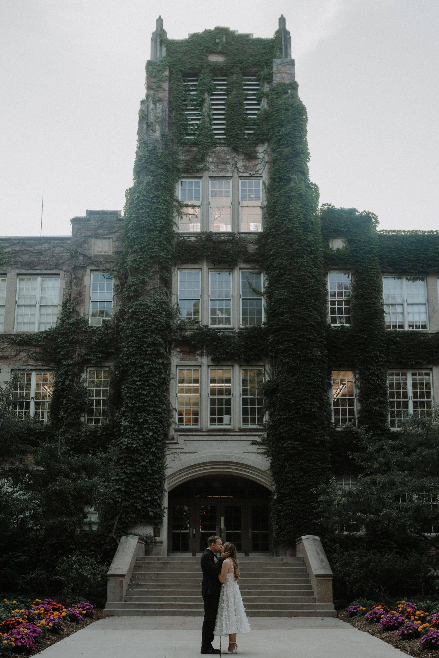 Elegant couple poses on steps of an ivy-covered Gothic university building with blooming flowers.