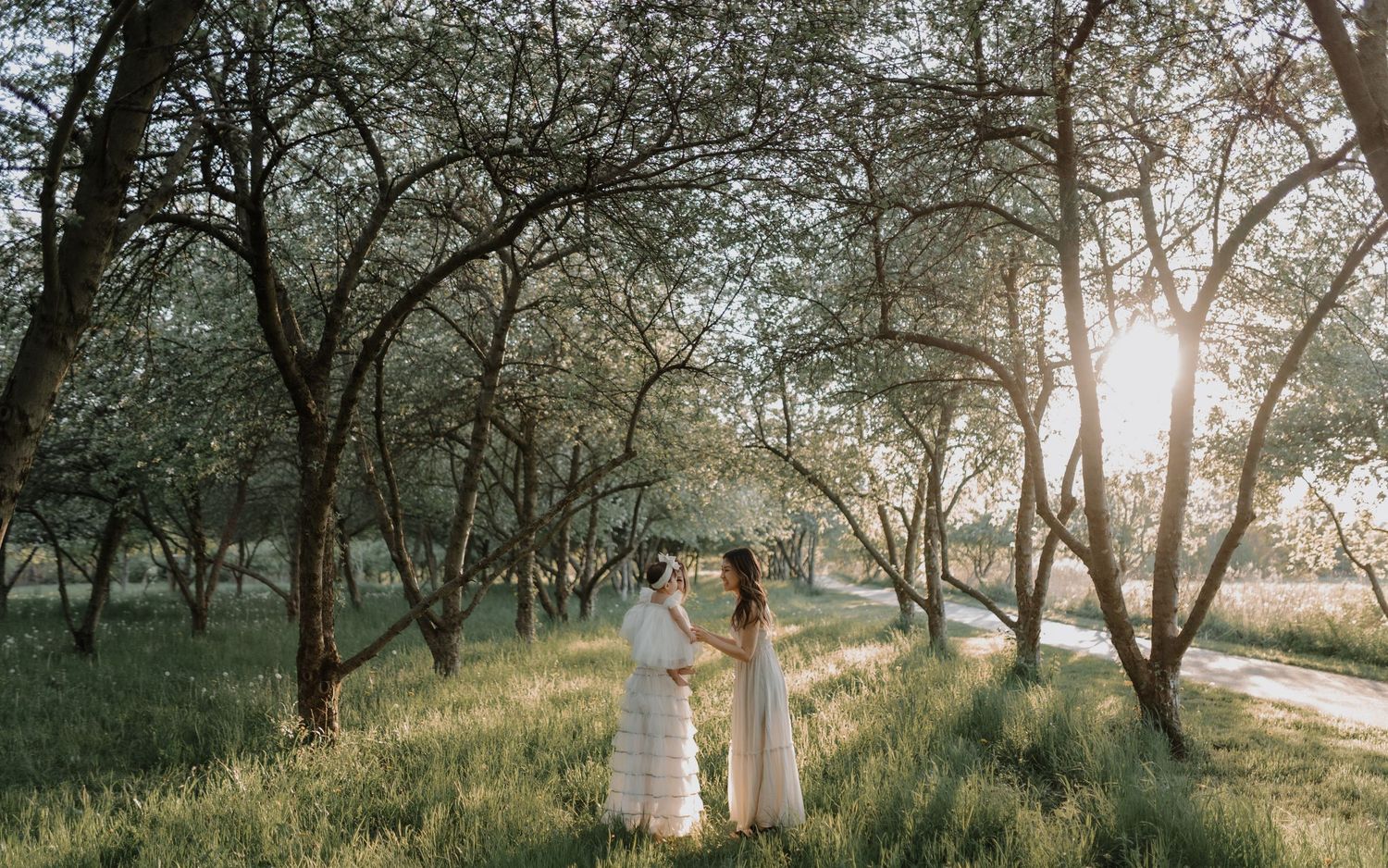 Two figures in flowing white dresses stand beneath blooming trees along a sunlit path during golden hour.