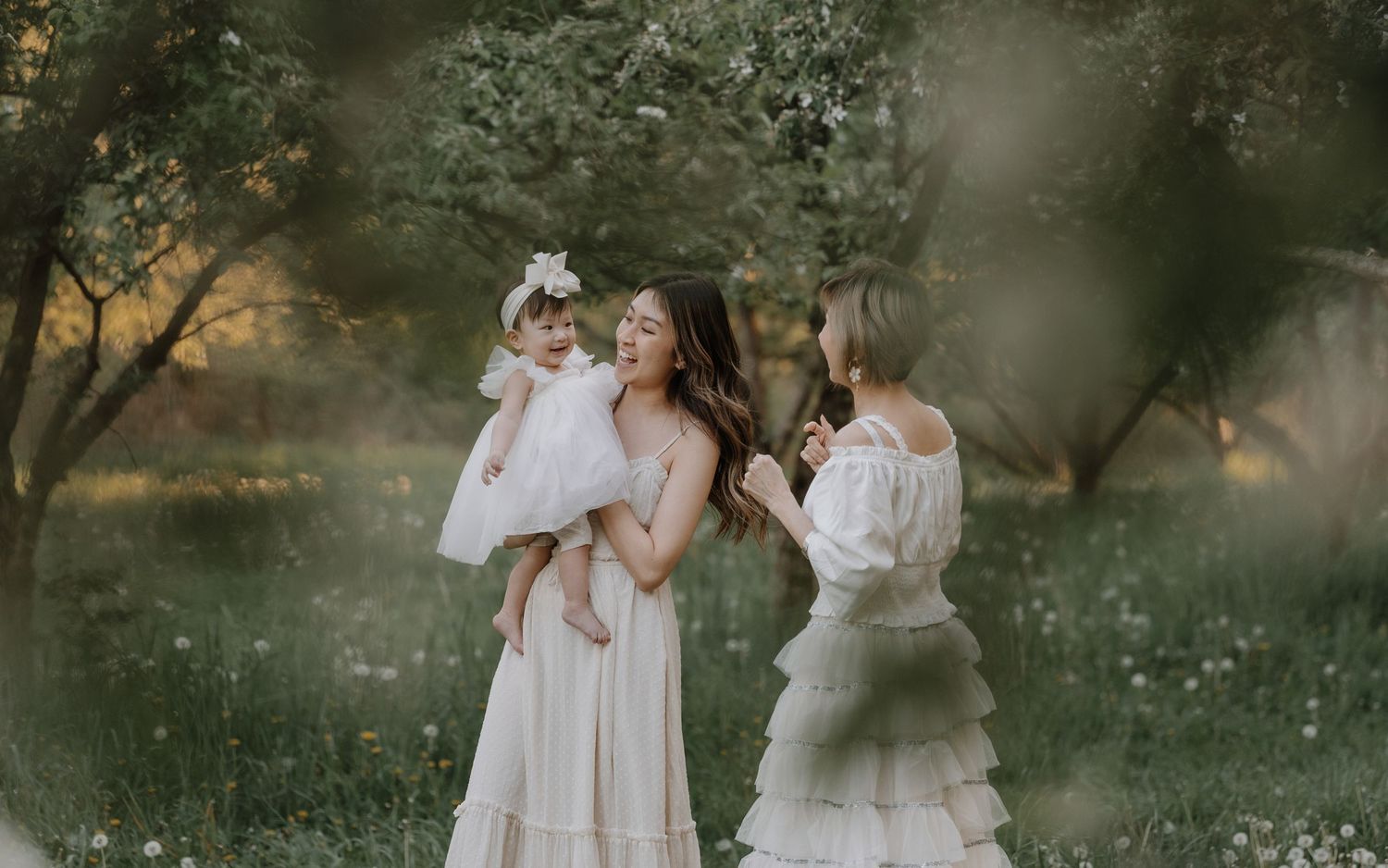 Three people in flowing white dresses stand together in a dreamy outdoor garden setting at golden hour.