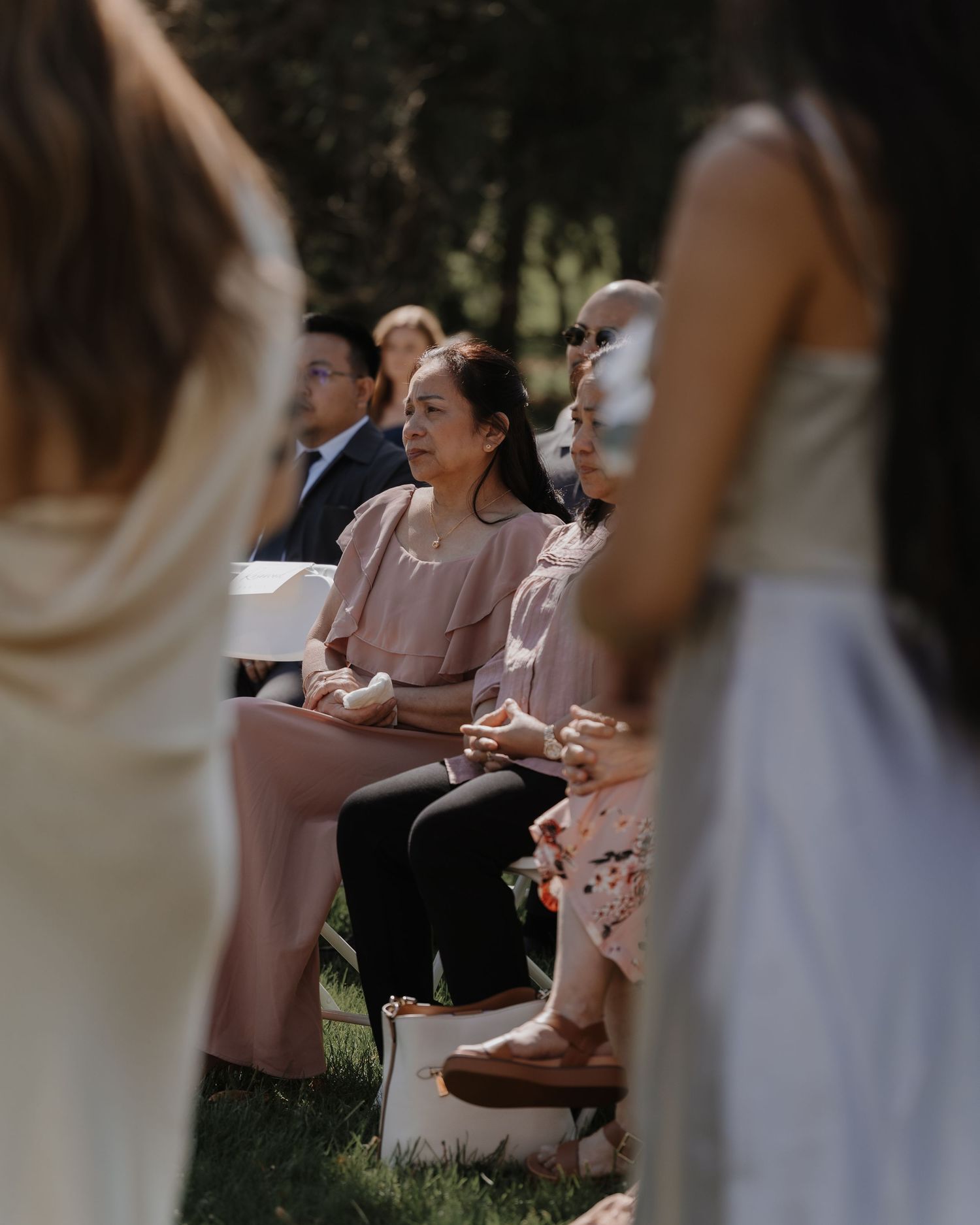 Guests seated at an outdoor wedding ceremony with white chairs and natural greenery in the background.