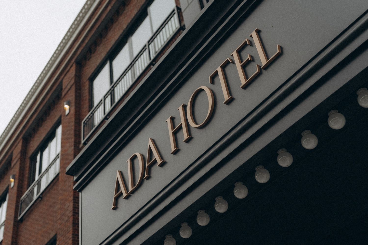 Exterior sign of the ADA Hotel with gold lettering on a dark facade, mounted on a classic brick building.