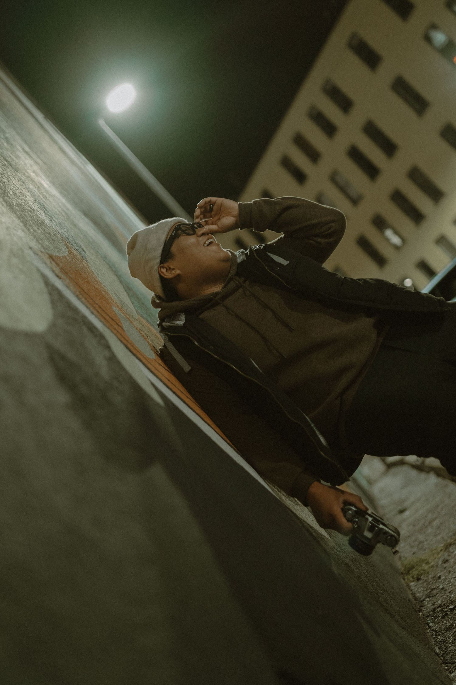 Young person in hoodie and cap leaning against wall at night with skateboard under urban street light.