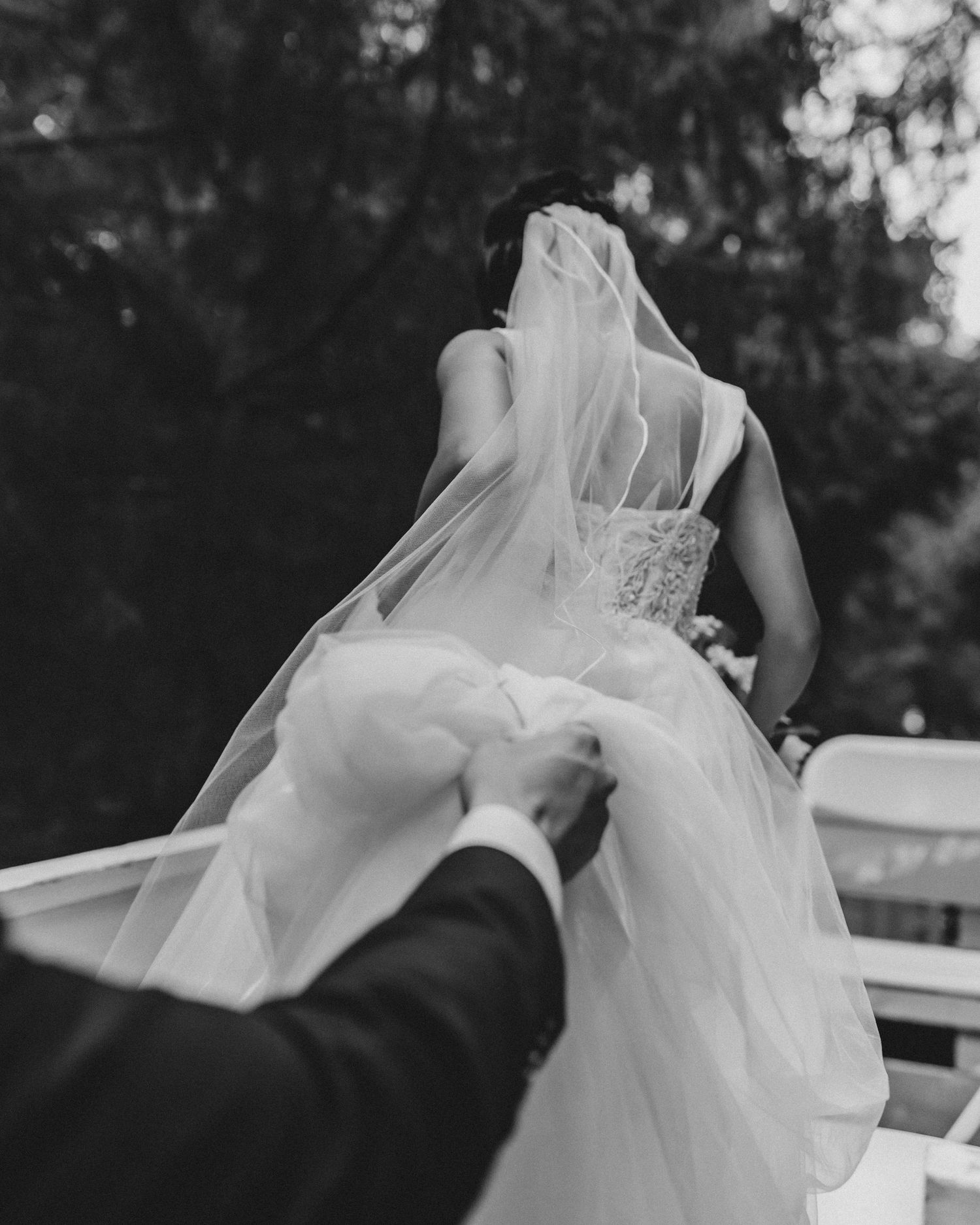 Black and white wedding photo showing a bride in a flowing white dress and veil being helped with her gown from behind.