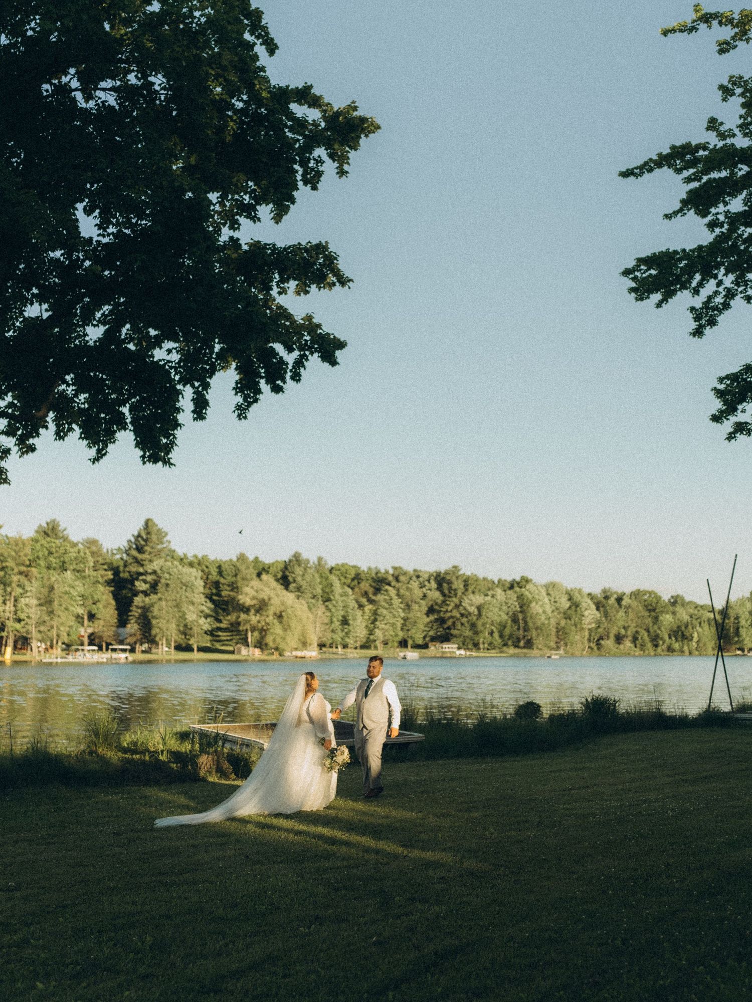 Bride and groom holding hands by a serene lake surrounded by lush green trees on a sunny wedding day located in Greenville, Michigan.