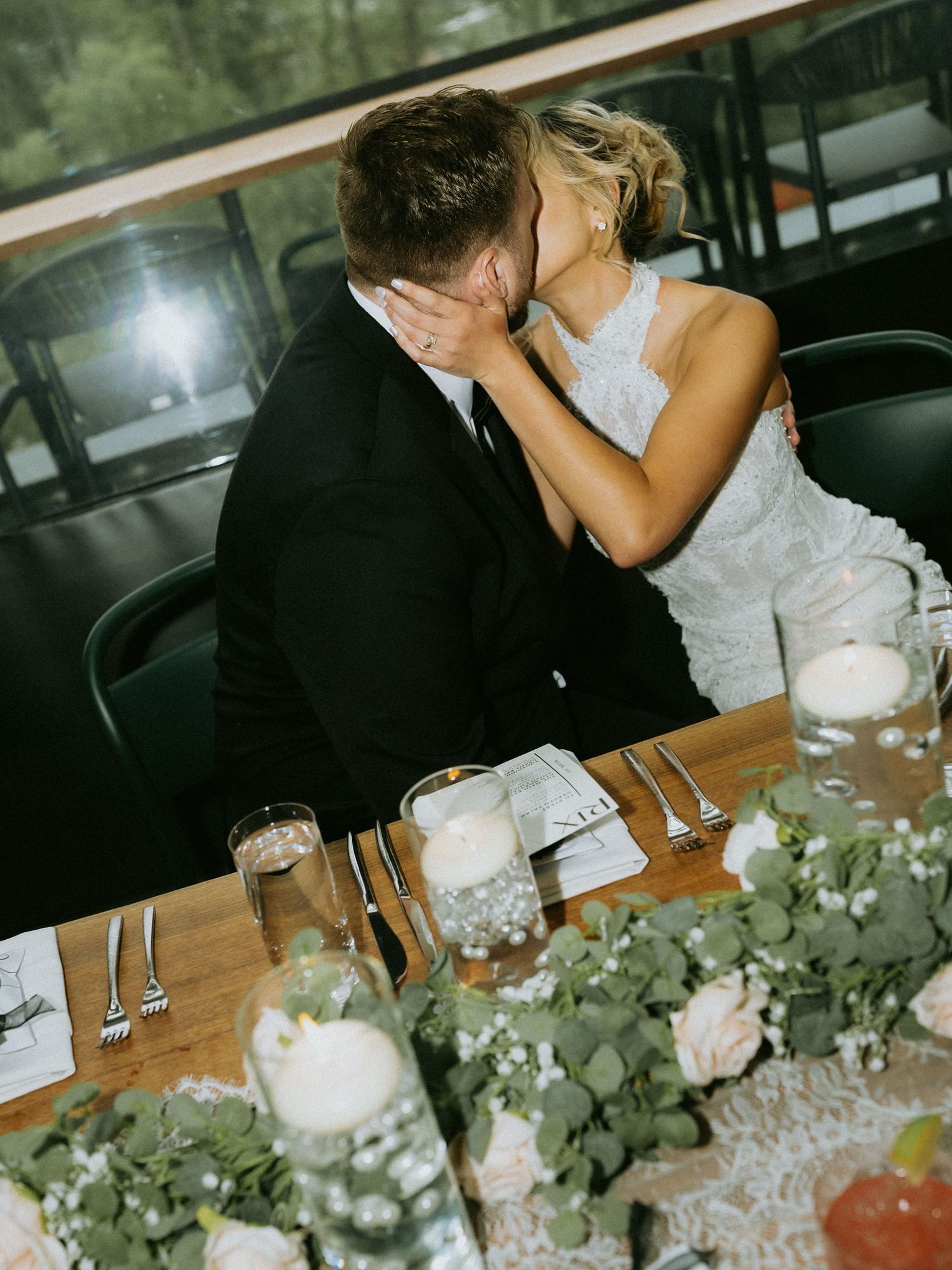 Bride and groom sharing a kiss at wedding reception table adorned with greenery, candles, and pink roses.