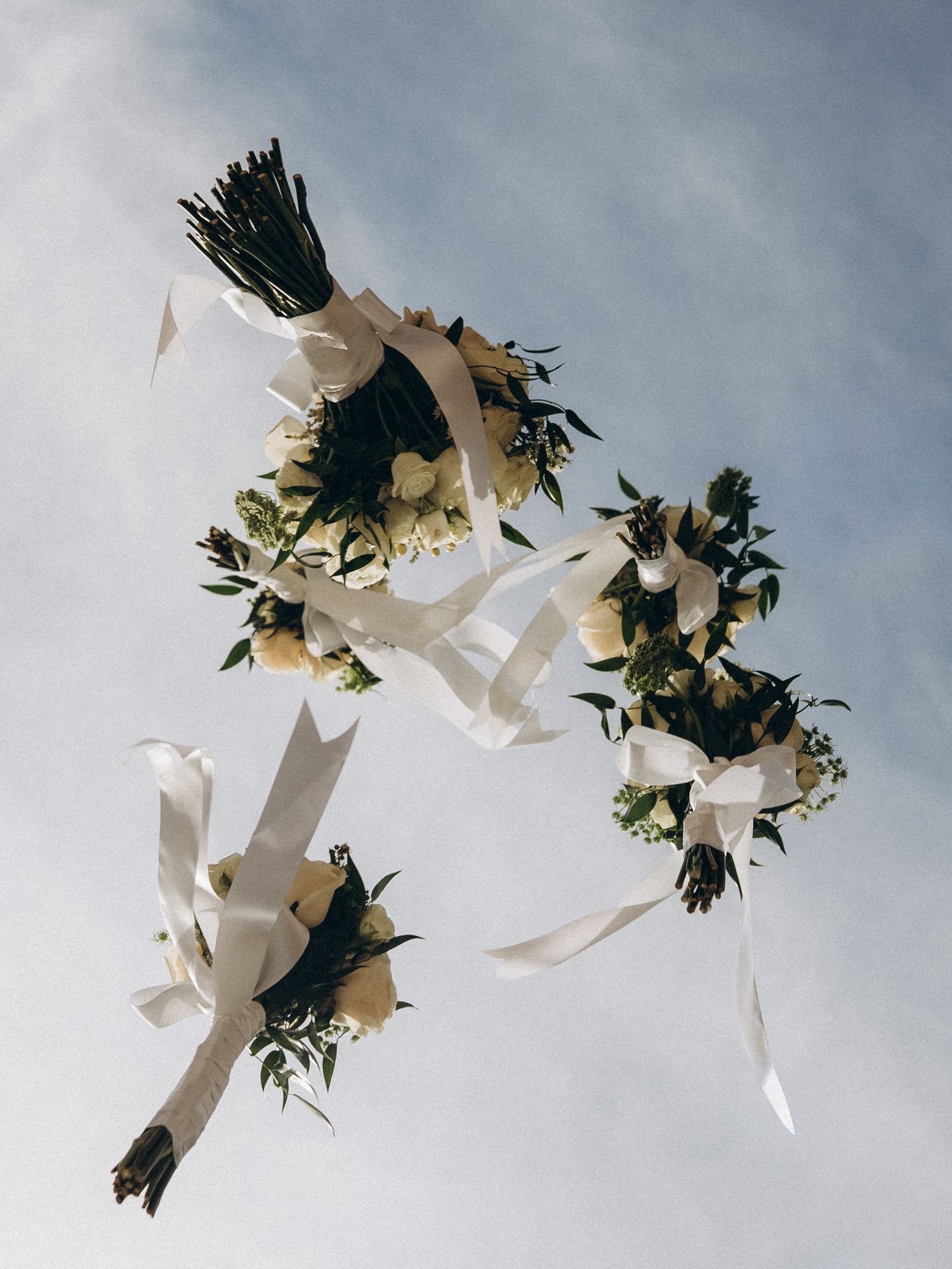 Three white floral wedding bouquets with ribbon streamers tossed against a blue sky.
