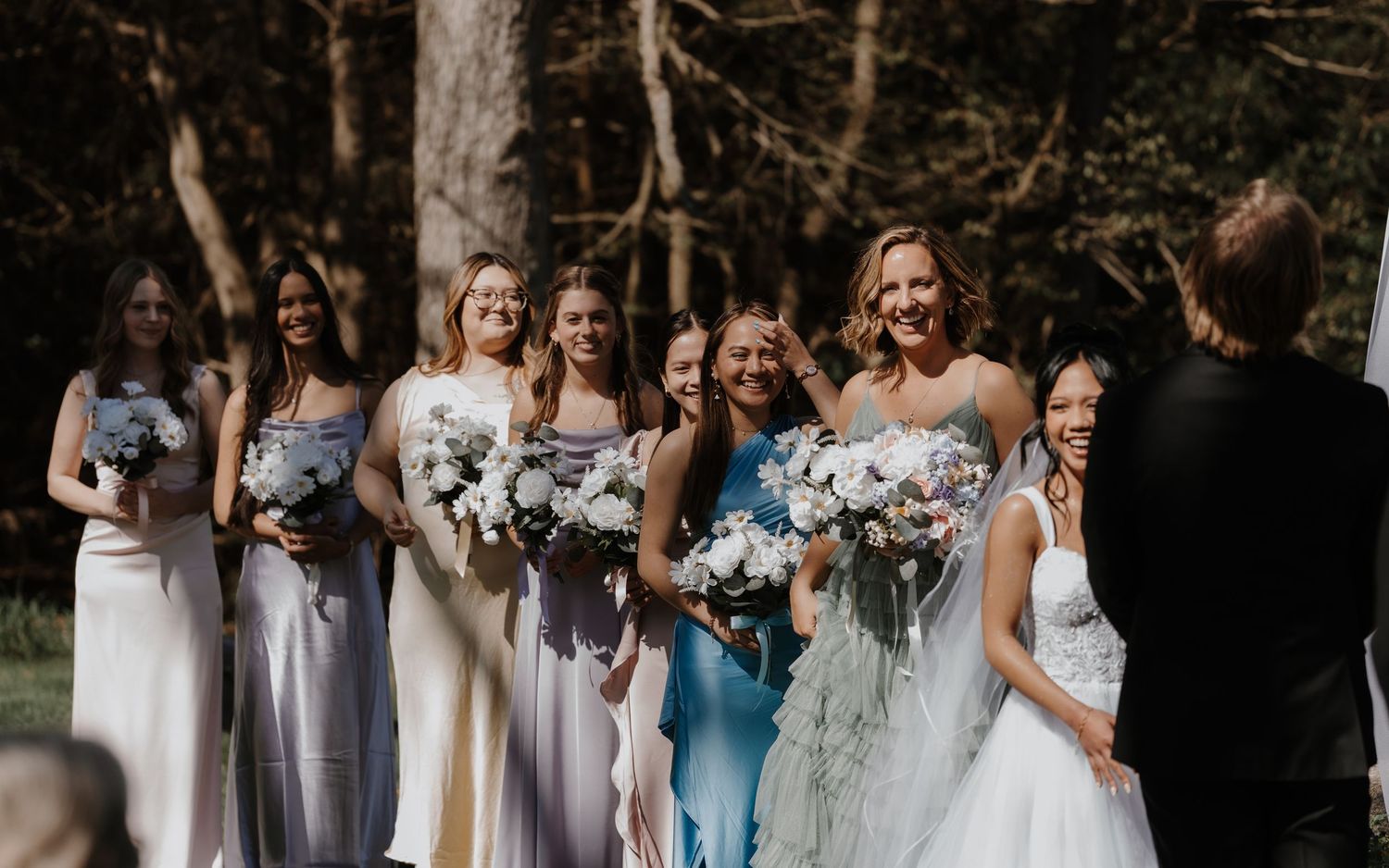 A wedding party in white dresses poses together in a wooded outdoor setting with blue floral bouquets.