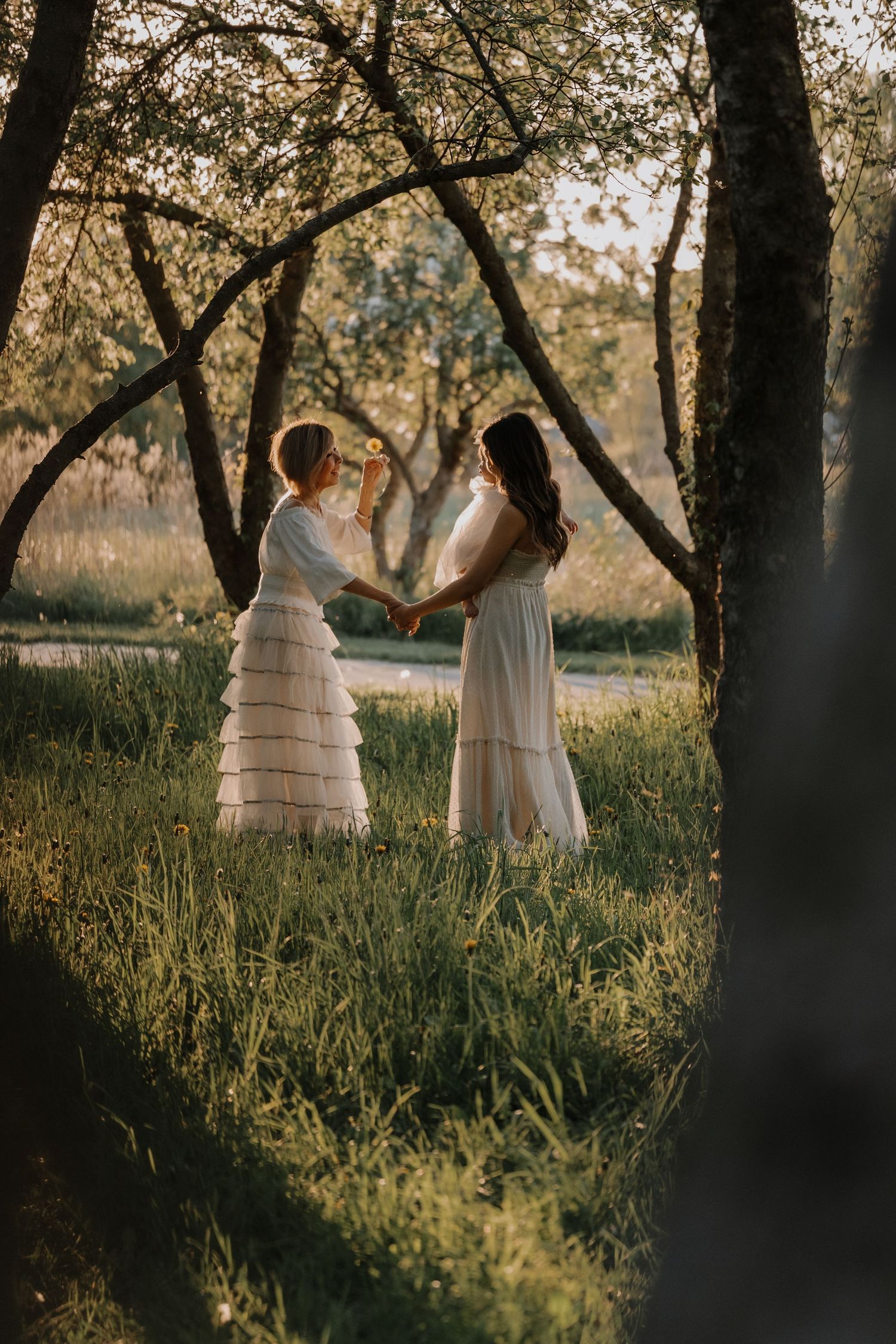Two people in white dresses hold hands while standing in a sunlit forest clearing at golden hour.