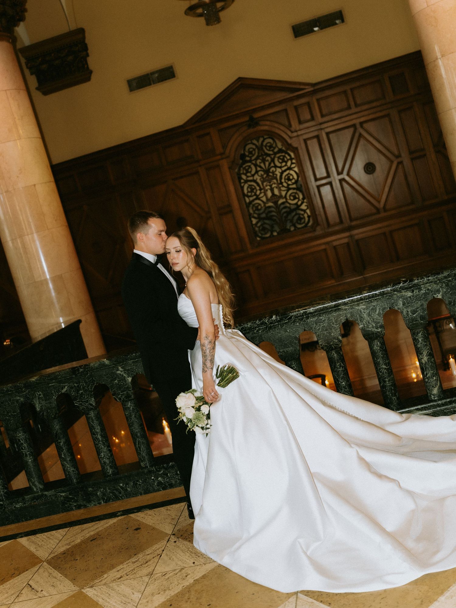Bride and groom pose elegantly in a grand hall, her white ballgown train flowing across marble floors.