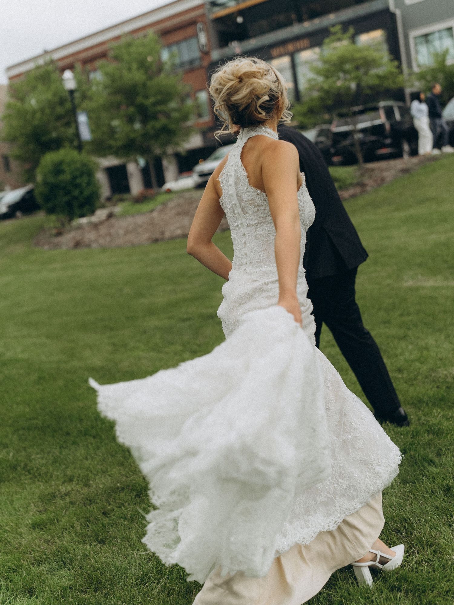 Bride in lace mermaid wedding gown and groom in black tuxedo walking on green lawn.