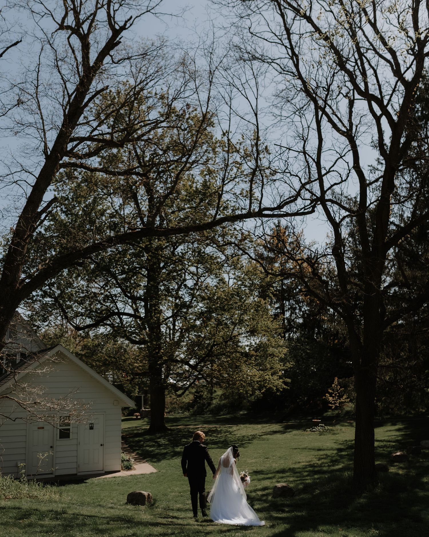 Two figures walk near a small white shed on a grassy lawn surrounded by bare trees in an artistic wedding photo.