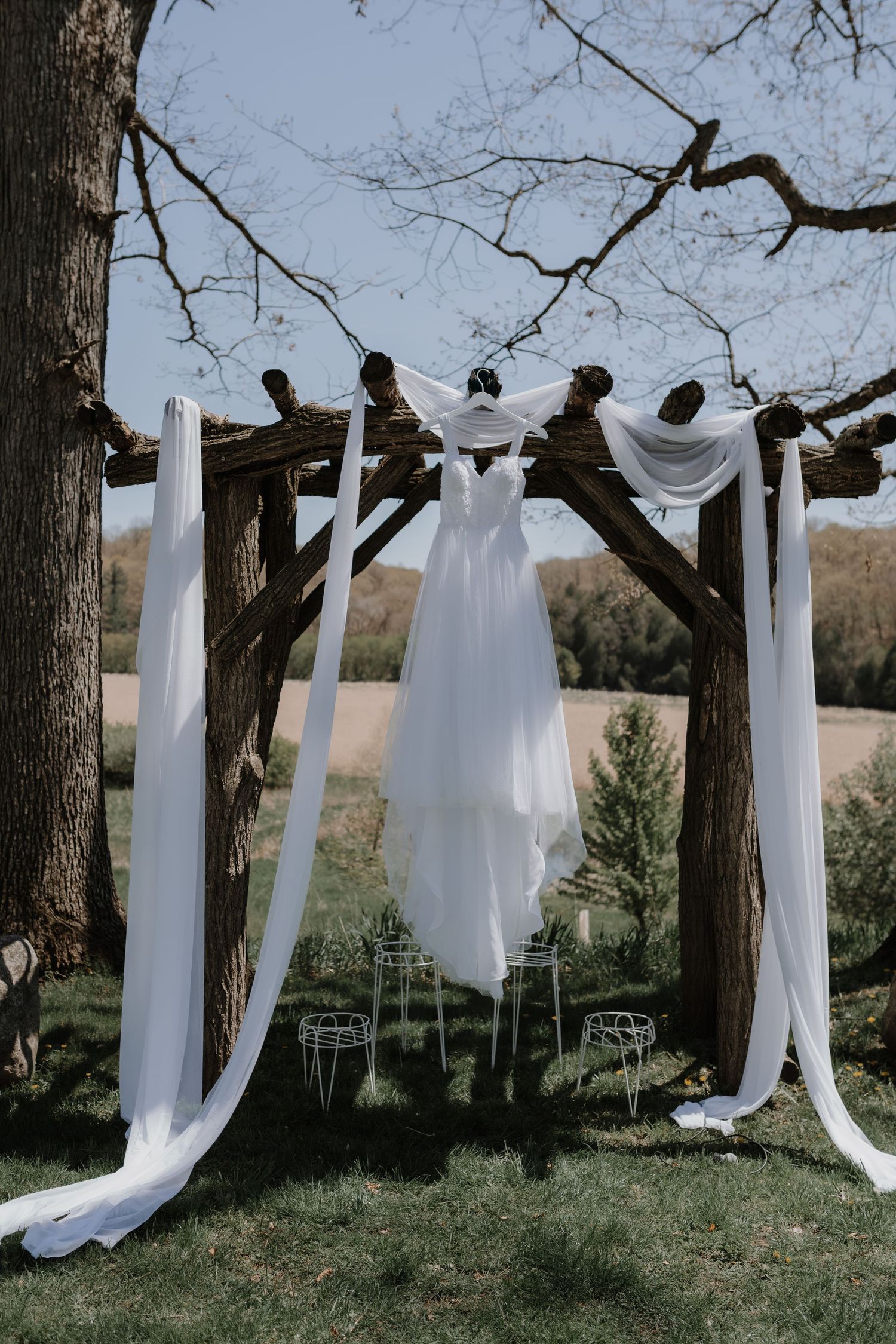 A white wedding dress and flowing fabric drapes hang elegantly from a rustic wooden arch against a bright blue sky.