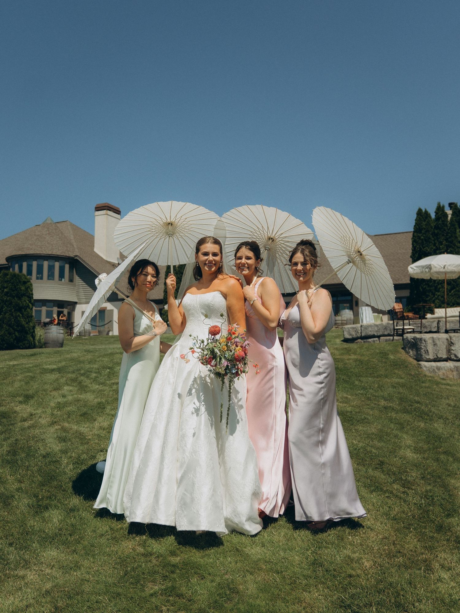 Bride and three bridesmaids holding white parasols pose outdoors on sunny day at elegant wedding venue.
