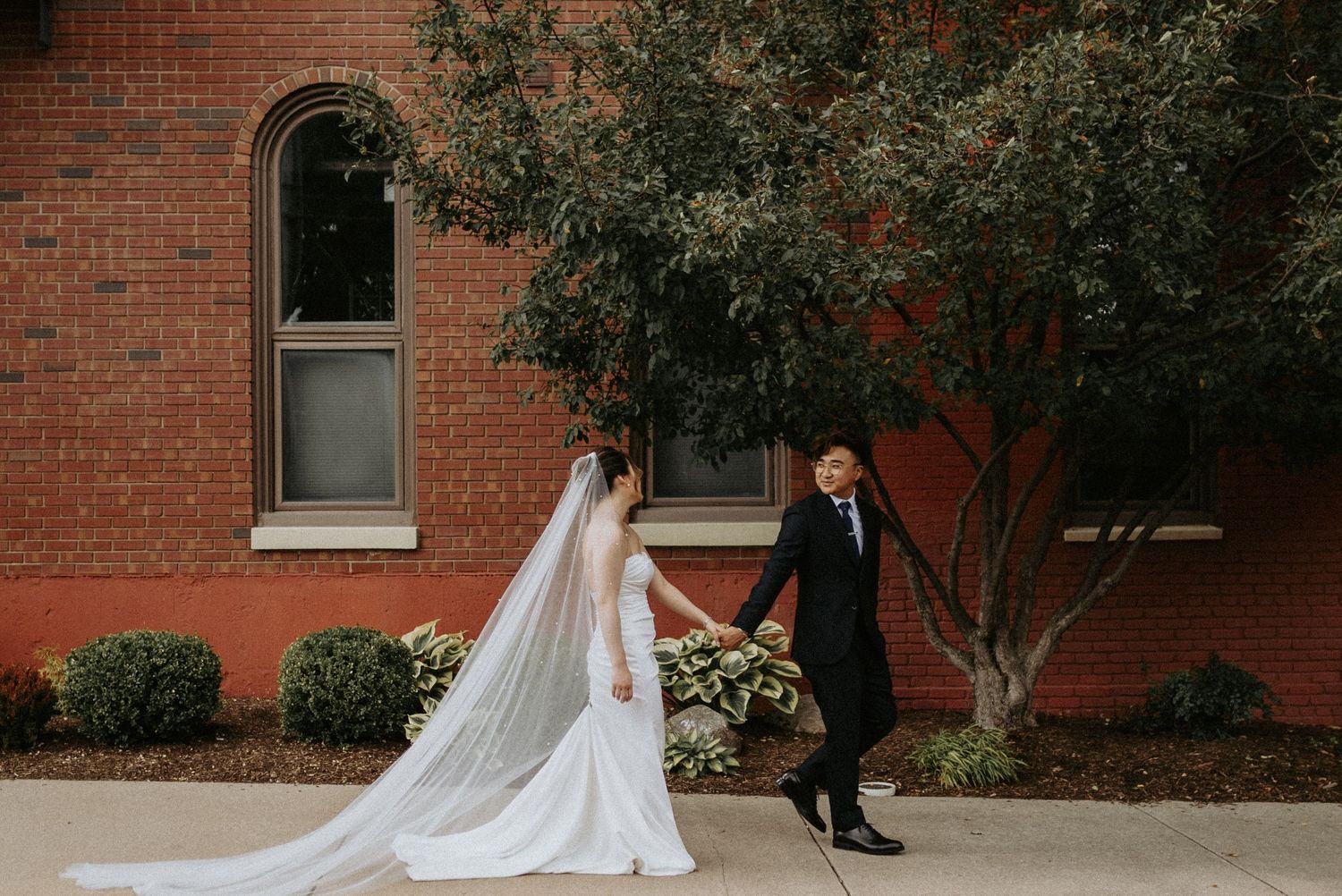 A couple in wedding attire walks together by a red brick building with a flowing long white veil trailing behind.