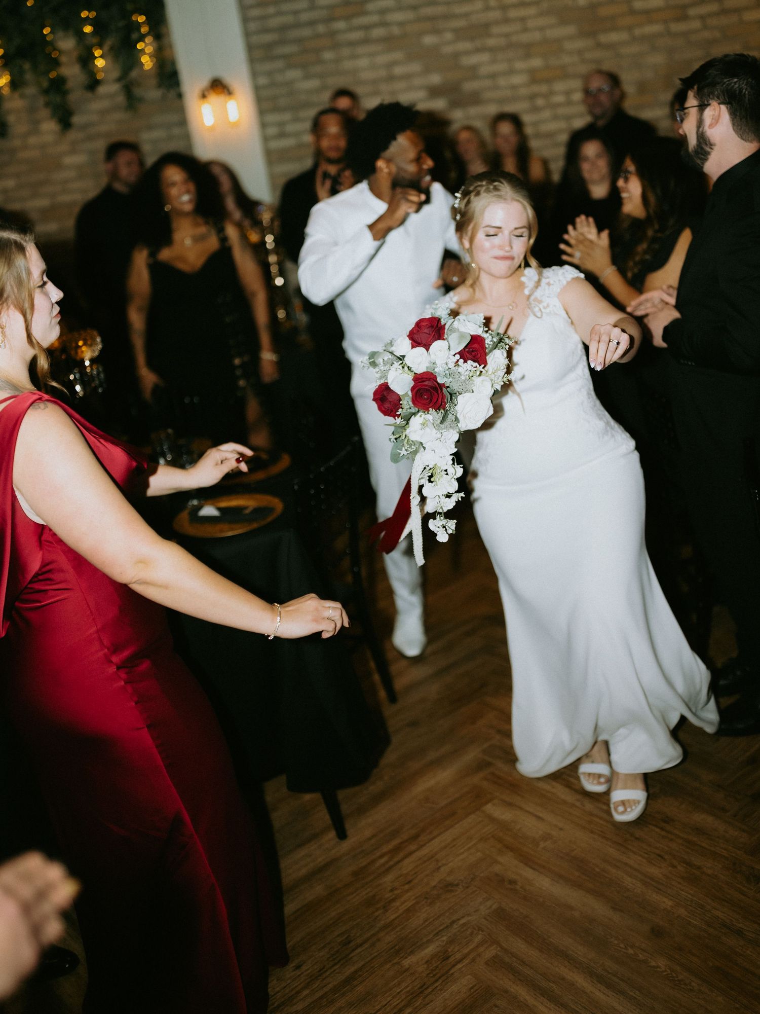 Bride in white gown holds red and white bouquet while dancing with guests at elegant wedding reception.