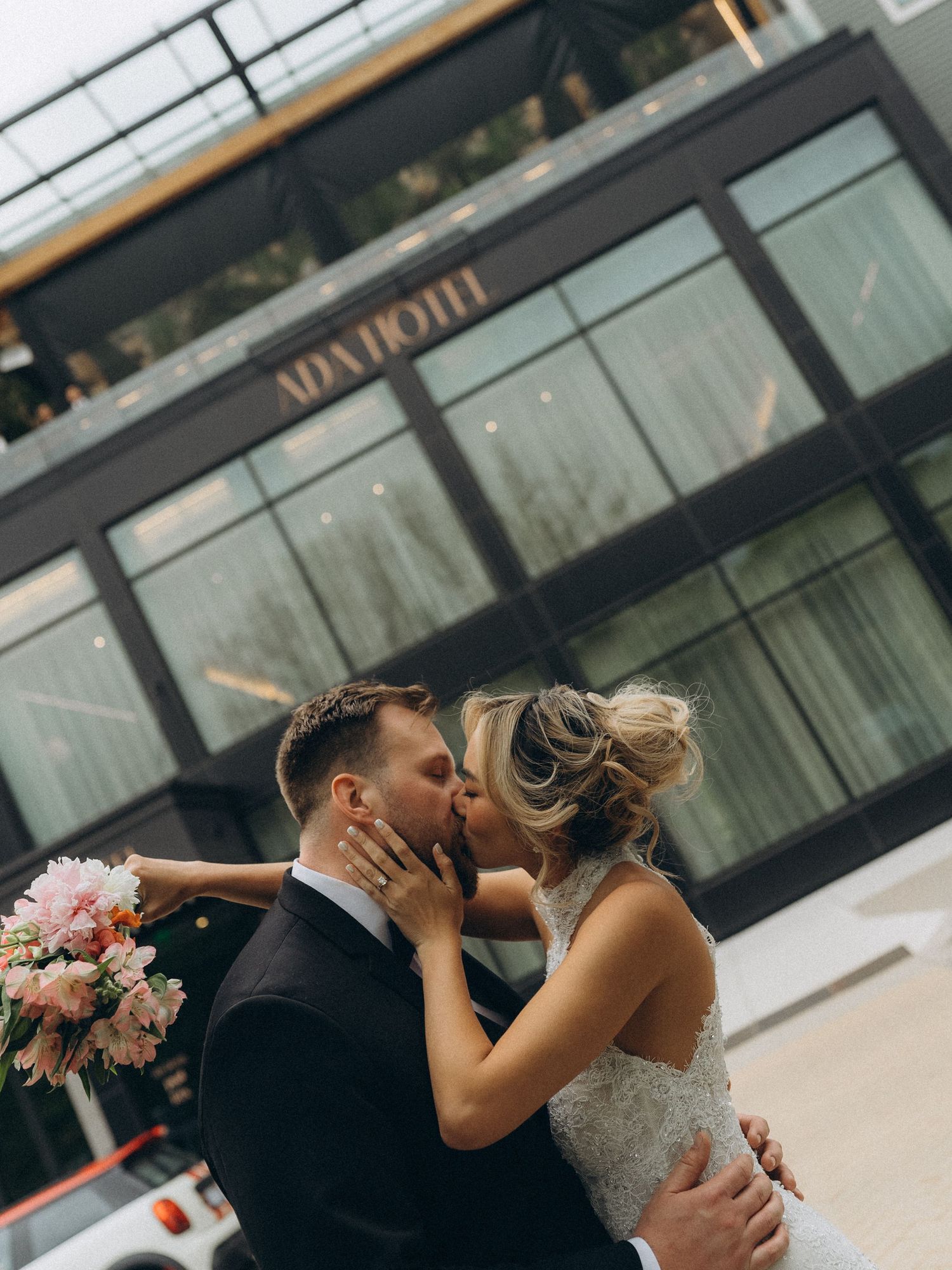 Newlywed couple sharing a romantic kiss outside the Ada Hotel, bride holding pink bouquet in elegant wedding attire.