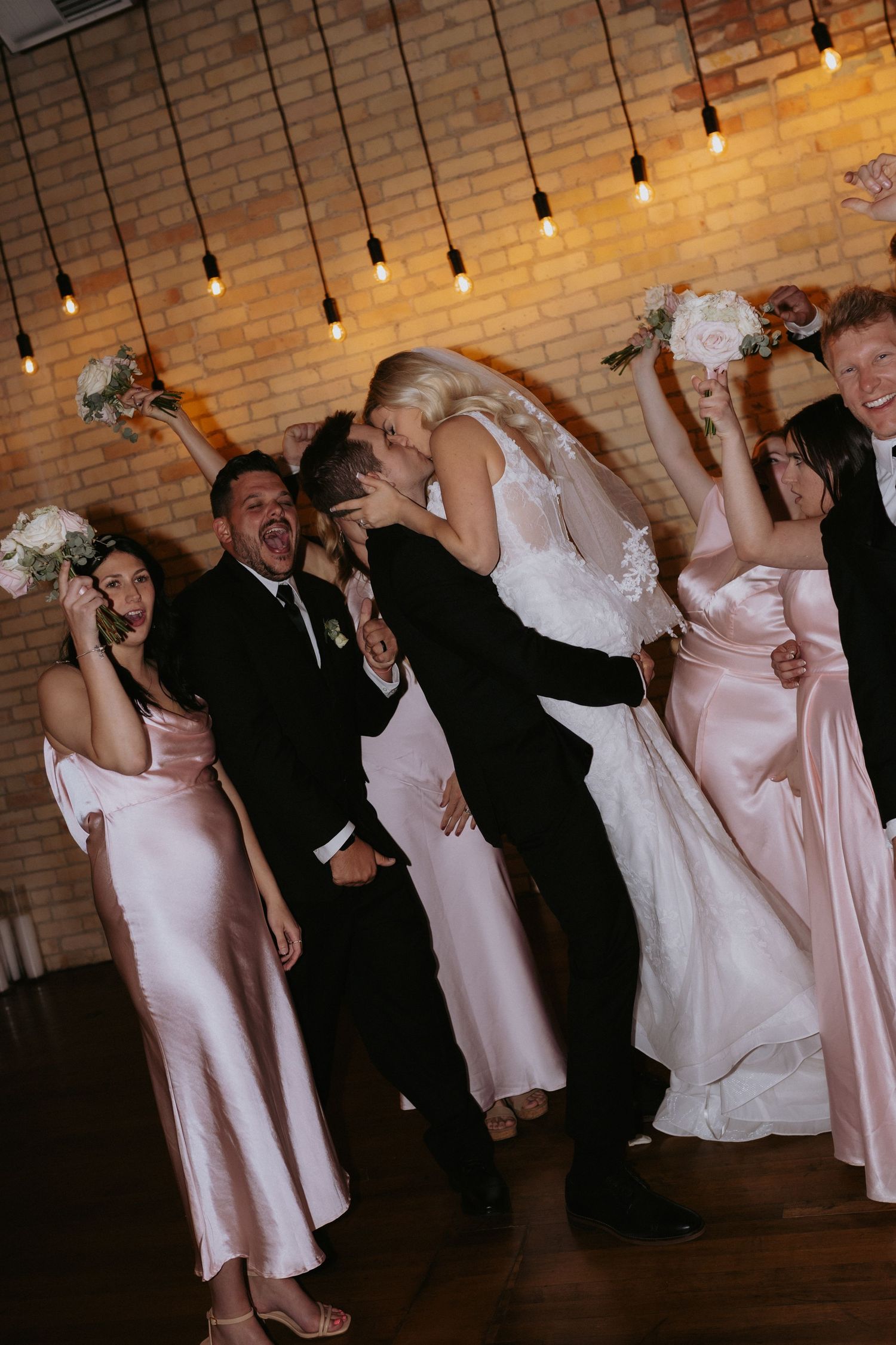 A group of wedding guests dance joyfully during reception festivities under string lights and brick walls.