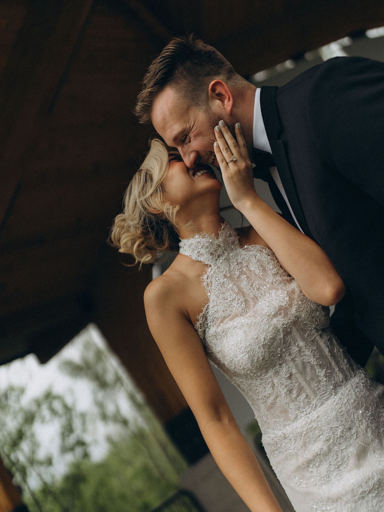 A bride and groom share a romantic kiss, she in lace gown, he in black tuxedo, at an outdoor wedding venue.