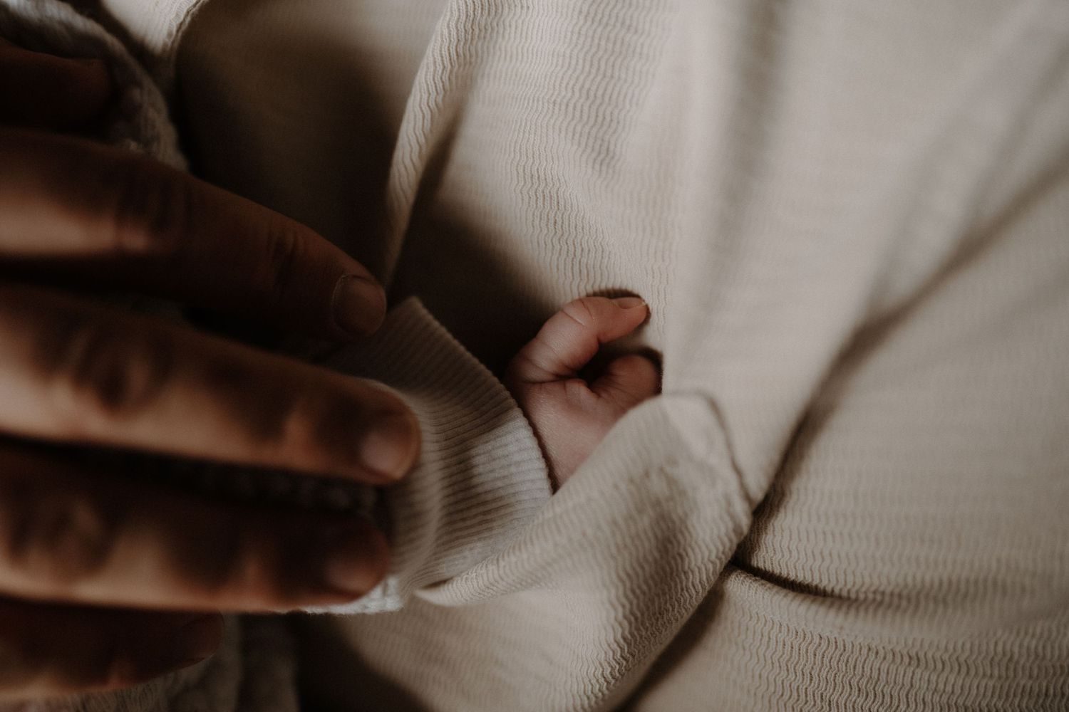 Close up view of a tiny newborn hand being gently held against soft white fabric in moody lighting.