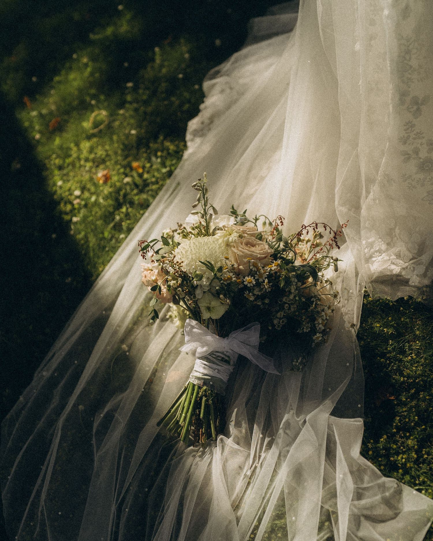 Bridal bouquet of white and blush flowers resting on a flowing wedding veil draped over lush green grass.