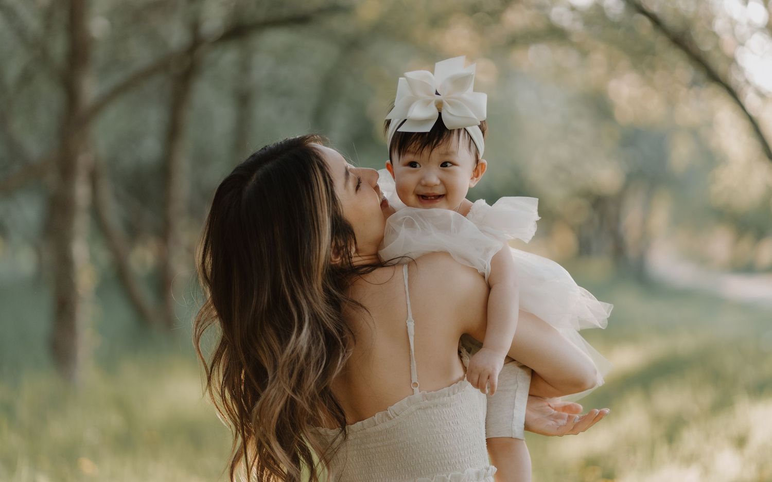 A mother and baby share tender moments embracing outdoors with sunlight filtering through trees in the background.