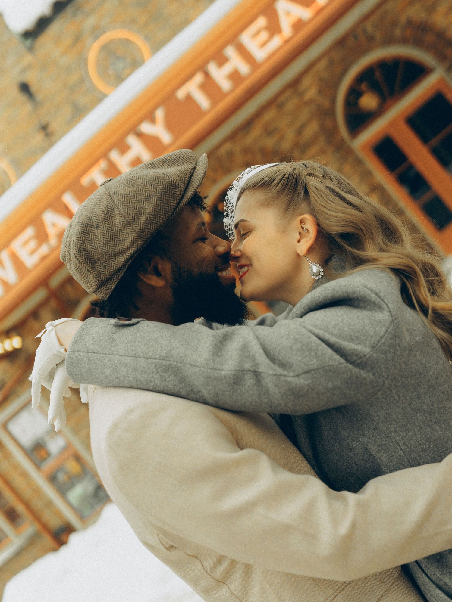 A couple sharing a romantic embrace outside the Wealthy Theatre, dressed in winter coats and a beret.