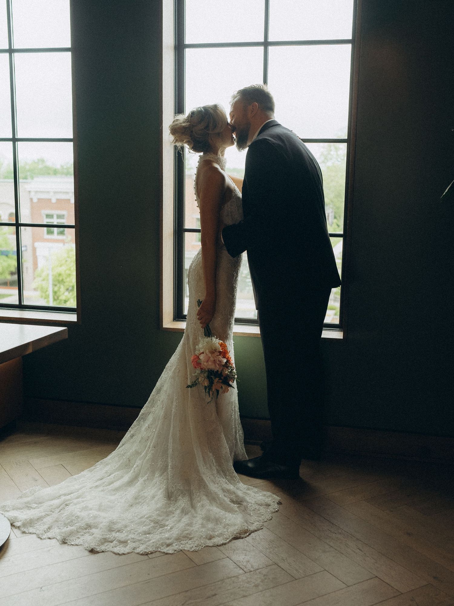 Bride and groom share a kiss by tall windows, her lace gown trailing elegantly on the floor.