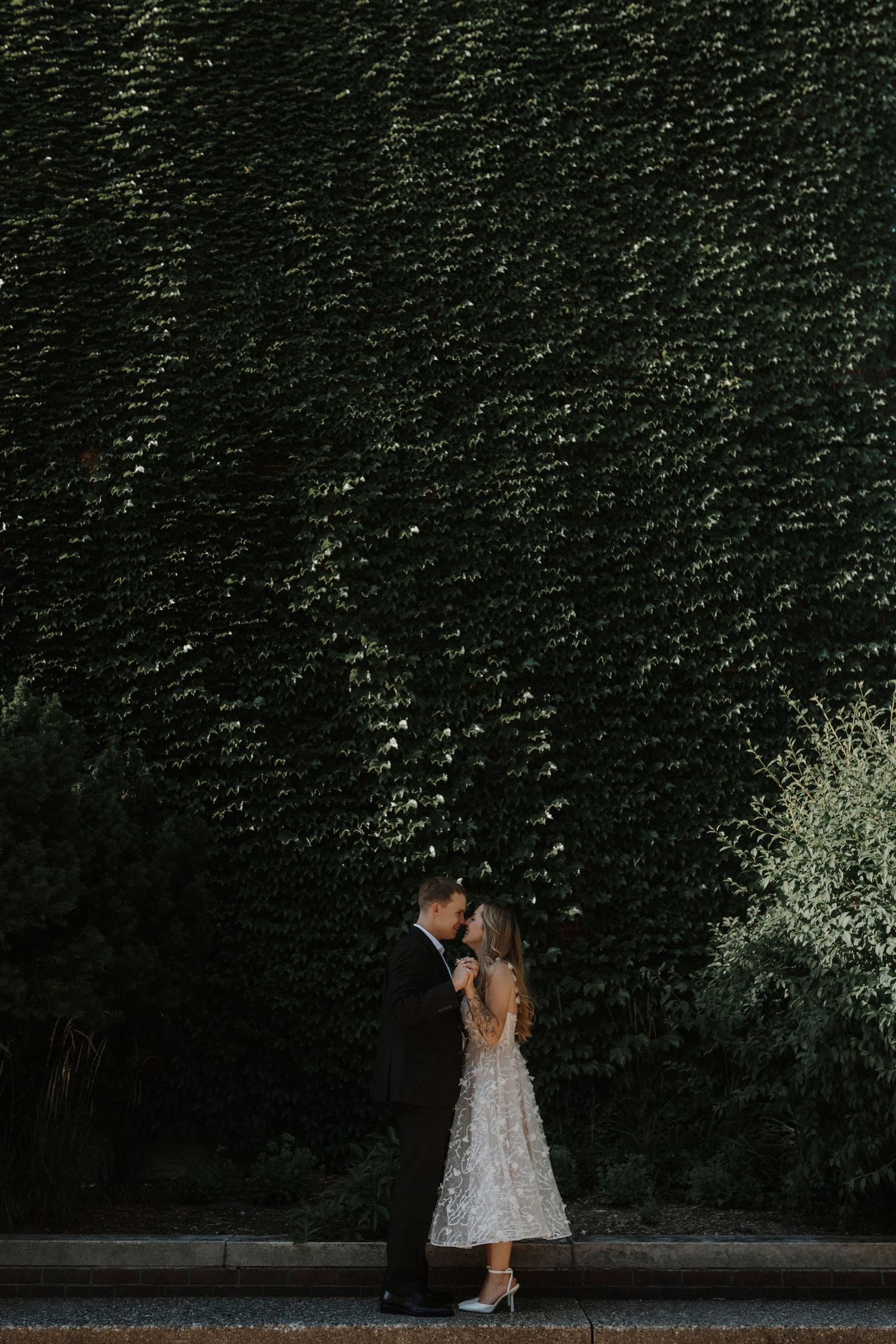A couple in wedding attire shares an intimate moment against a backdrop of lush green hedges in an outdoor garden setting.