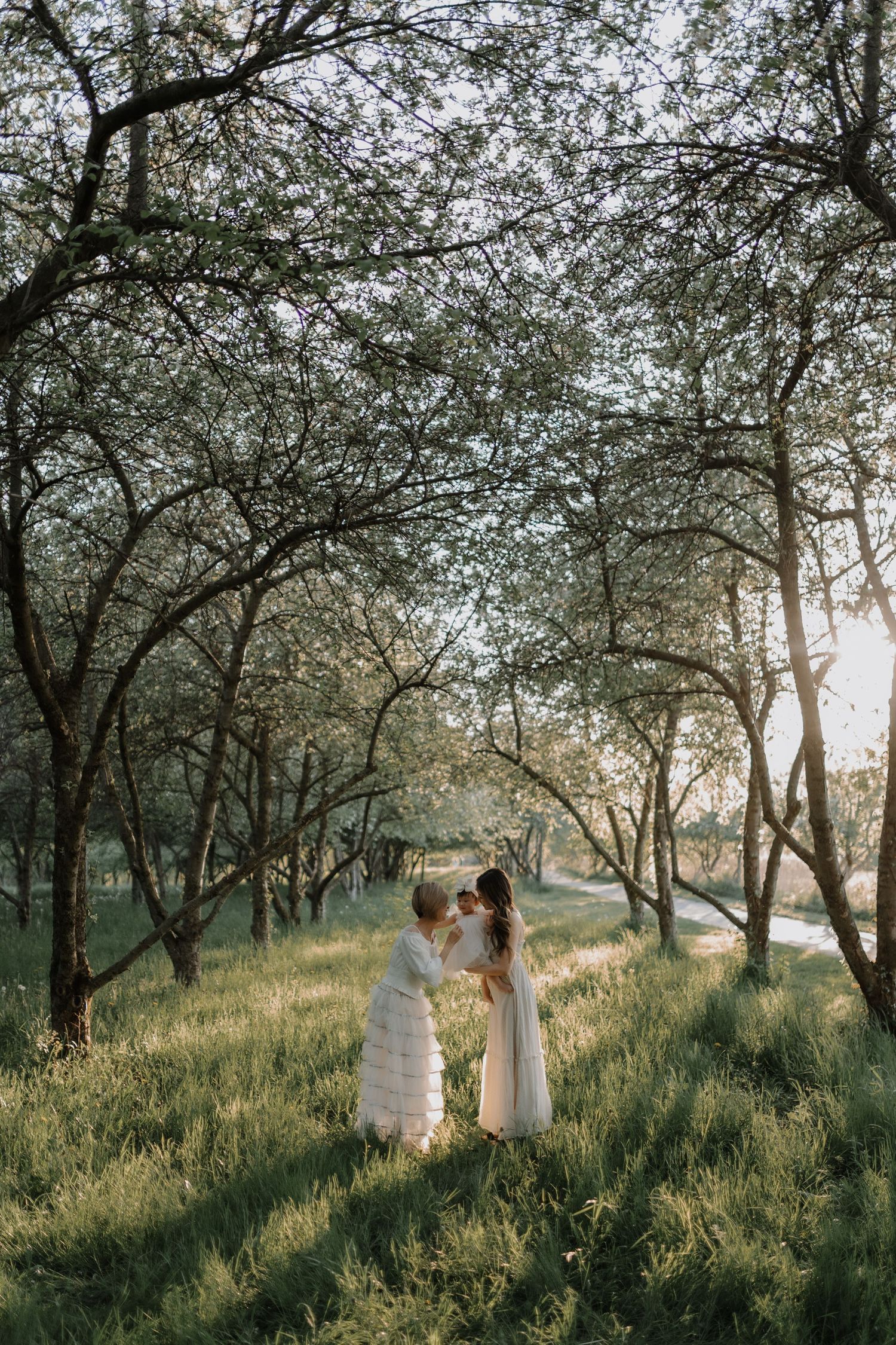 A couple in white clothing walks through a dreamy orchard with blossoming trees during golden hour.