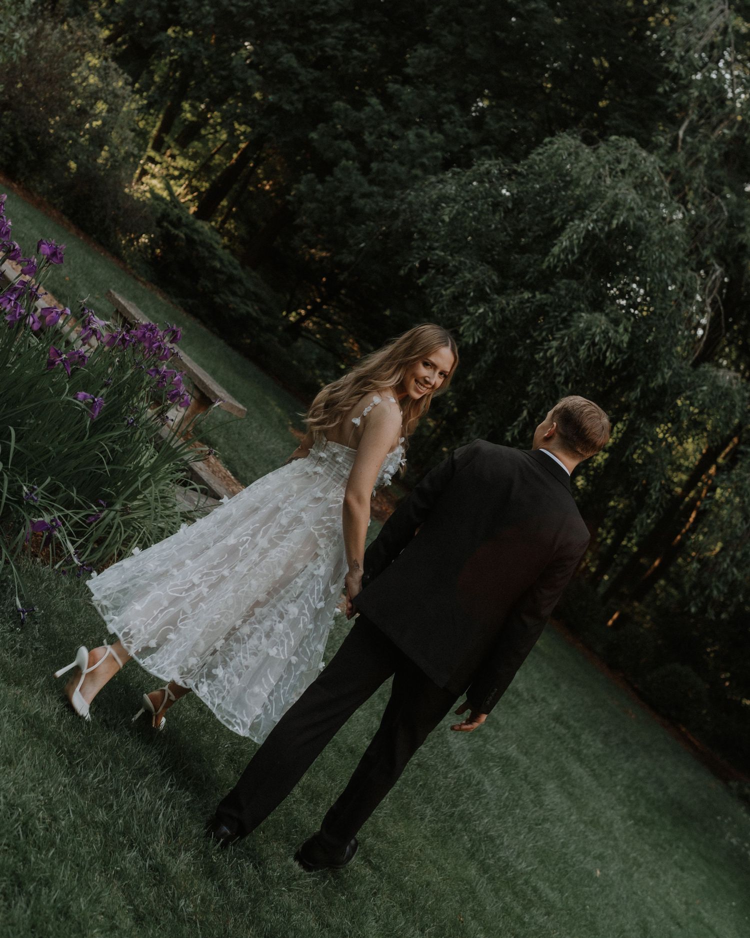 A couple in wedding attire stands on a green lawn with purple flowers and trees in the background during an outdoor photoshoot.