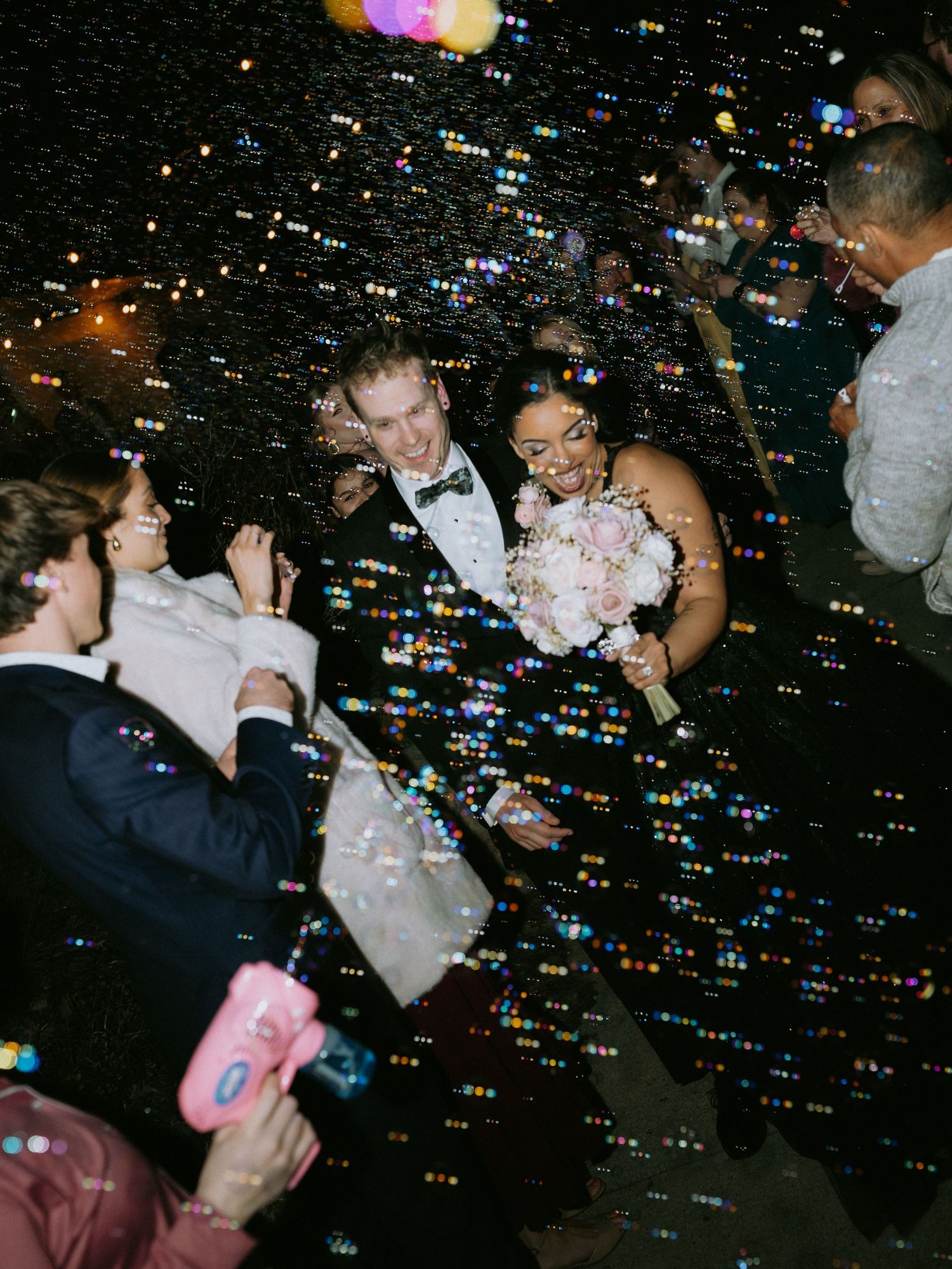 Bride and groom walk through bubble sendoff at night wedding reception surrounded by cheering guests.
