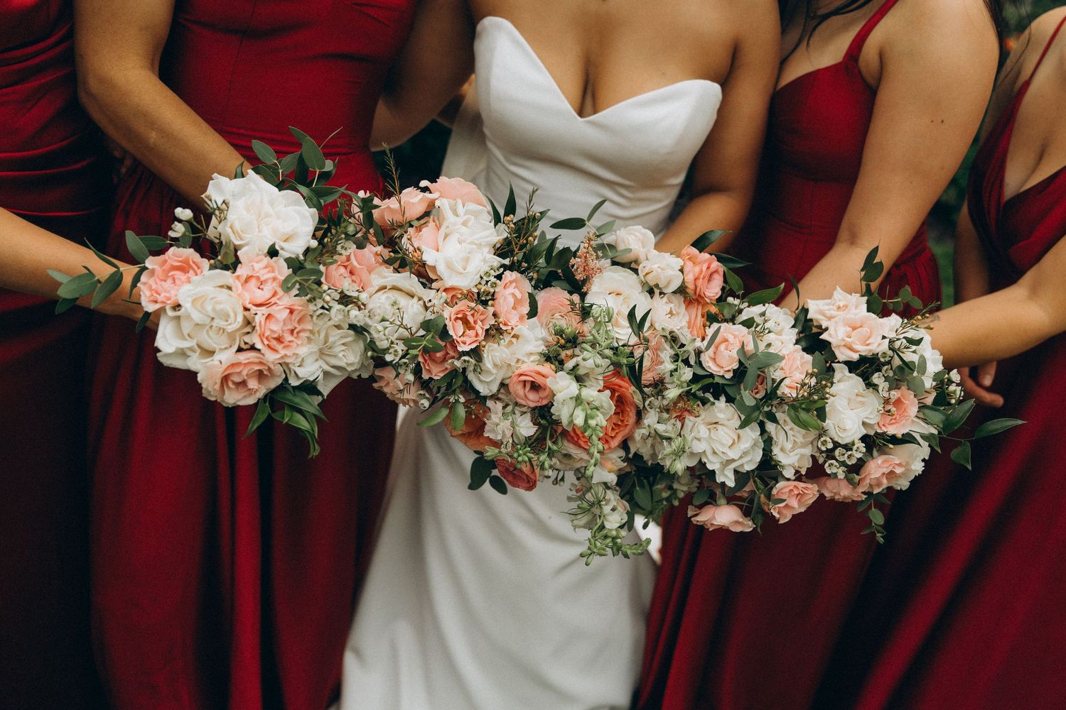 Bride in white gown and bridesmaids in red dresses holding lush pink and white floral bouquets at wedding.