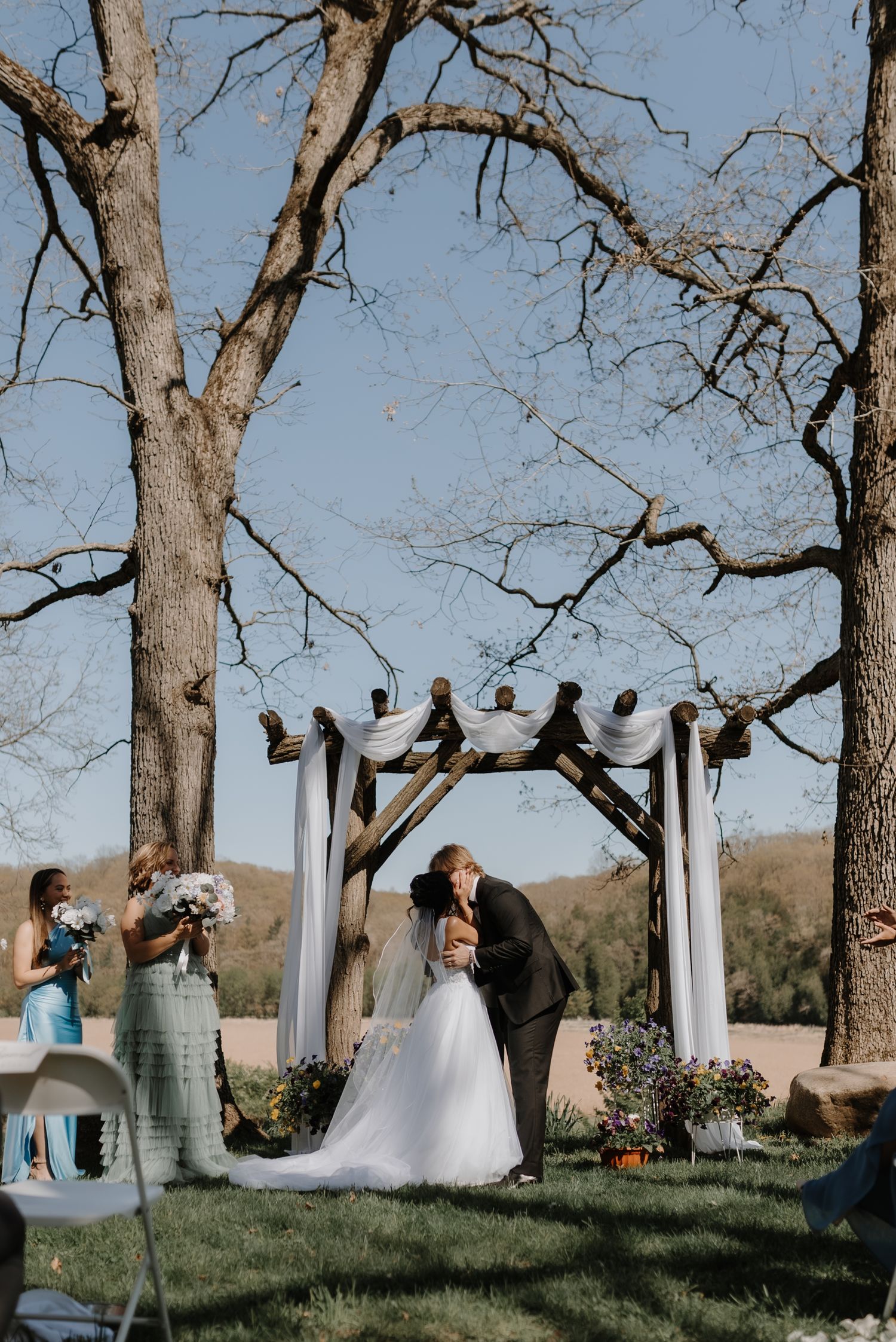 An outdoor wedding ceremony takes place under a white wooden arch between tall bare trees on a sunny day.