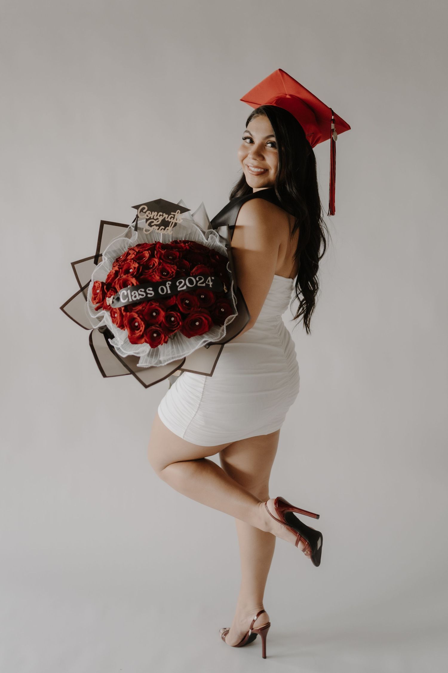 Graduate in white dress holds red rose bouquet while posing against grey background with red graduation cap.