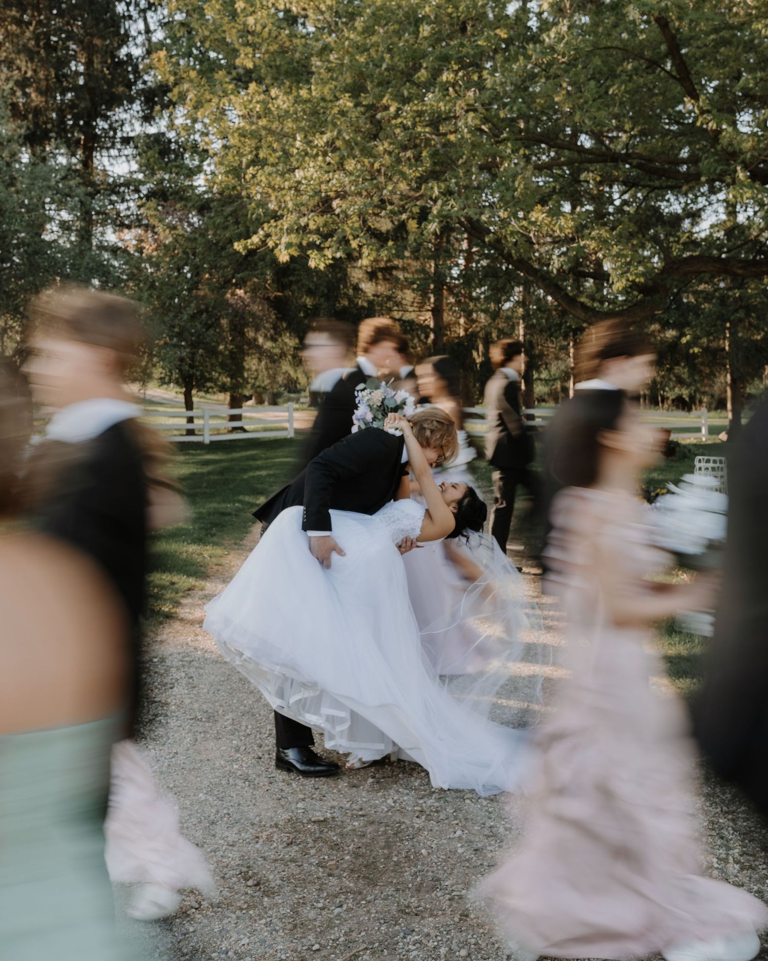 Motion-blurred wedding party members walk past as couple share romantic dip kiss on gravel path surrounded by autumn trees.
