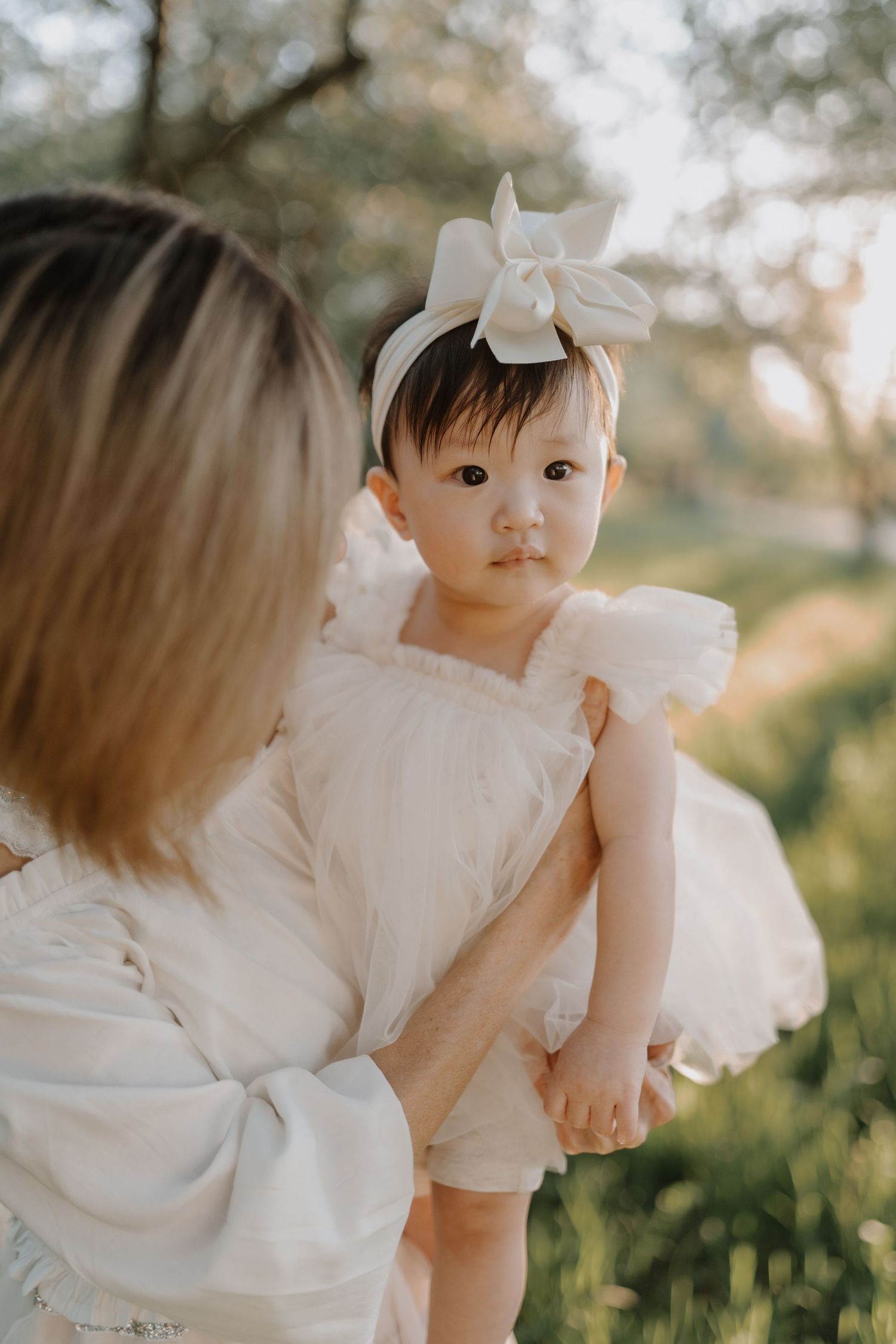 Child wearing white dress and bow against soft outdoor backdrop creates dreamy portrait composition.