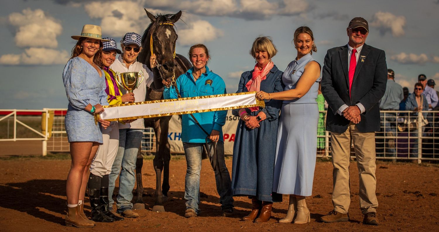 Group of people posing with a racehorse and winner's sash at a country horse racing event at sunset.