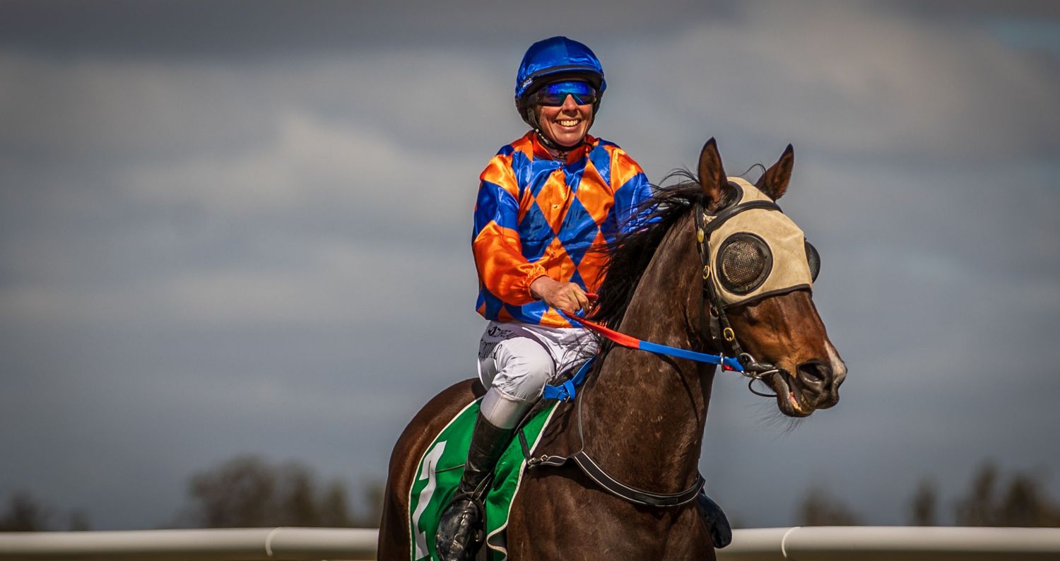 A smiling jockey in colorful orange and blue silks rides a chestnut horse on a racetrack under cloudy skies.