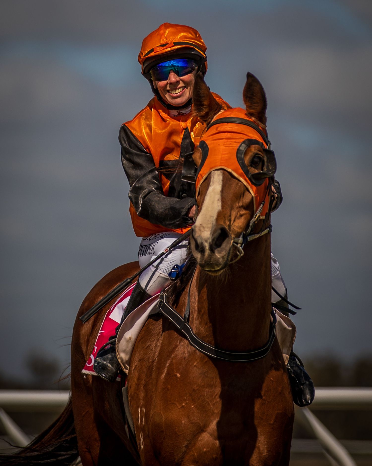 Smiling jockey in orange silks and helmet rides a chestnut horse on a sunny racetrack.