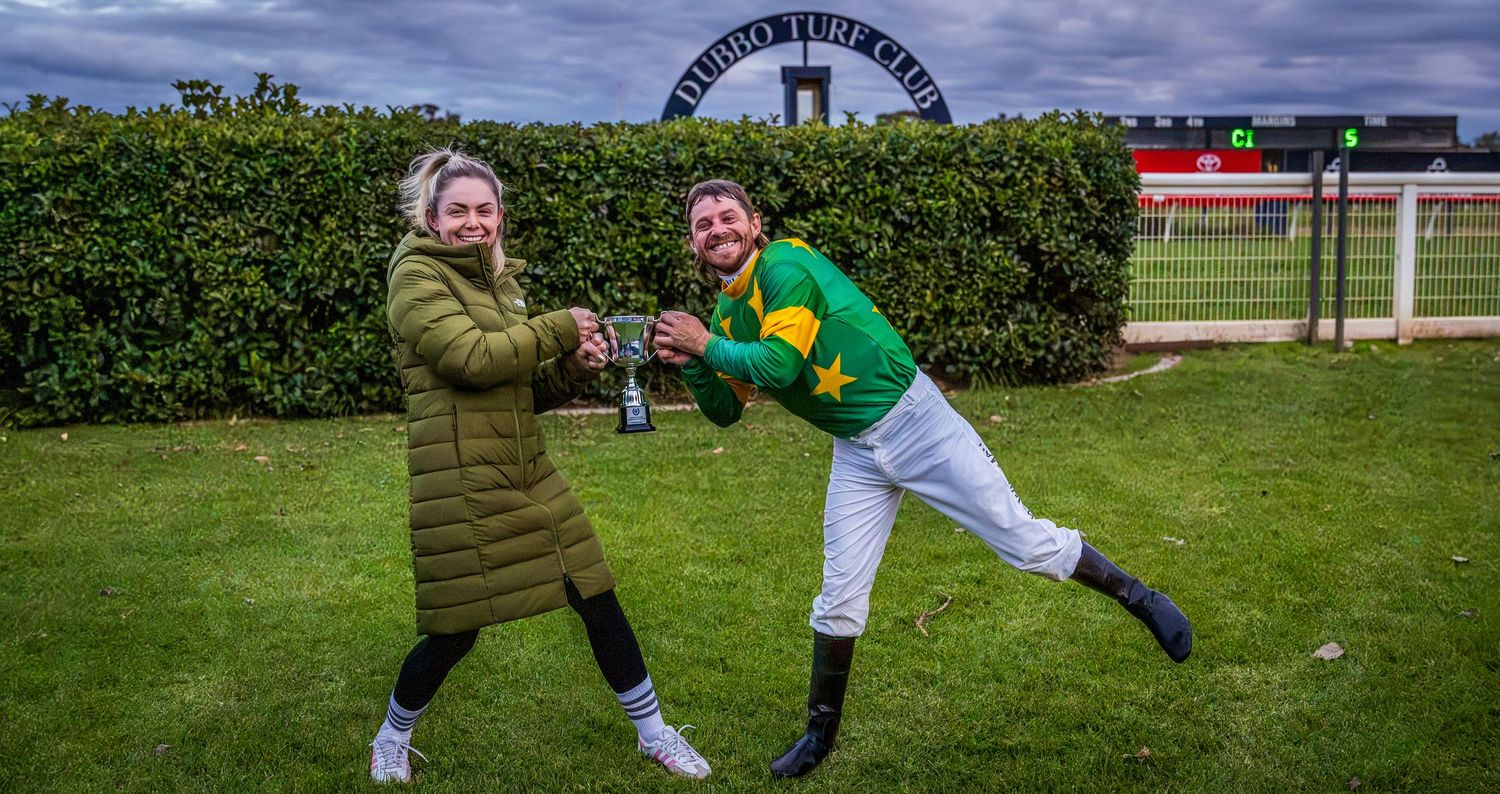 A jockey in green and yellow silks celebrates with a woman holding trophies at a turf club racecourse.