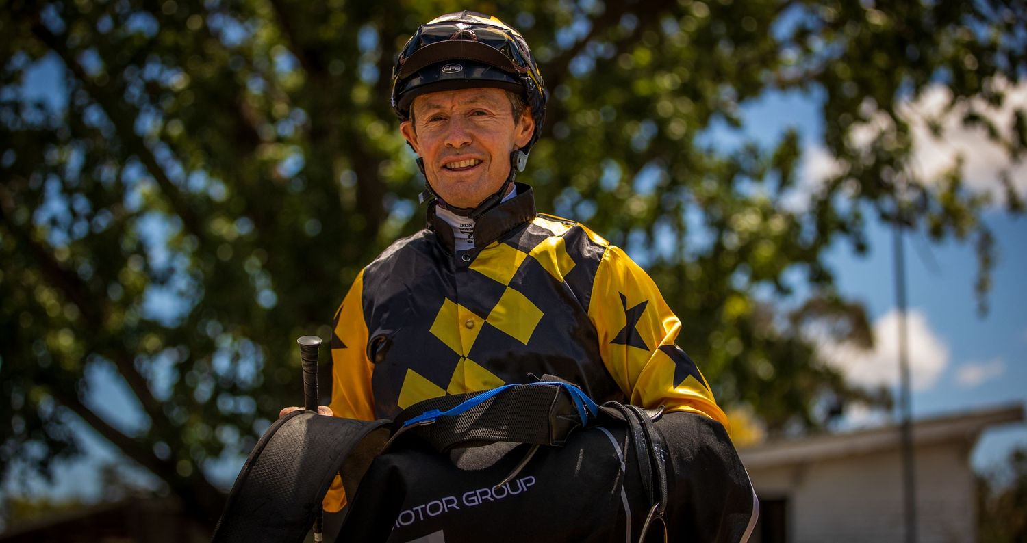 Smiling jockey in black and yellow checkered silks and helmet posing outdoors after a horse race.