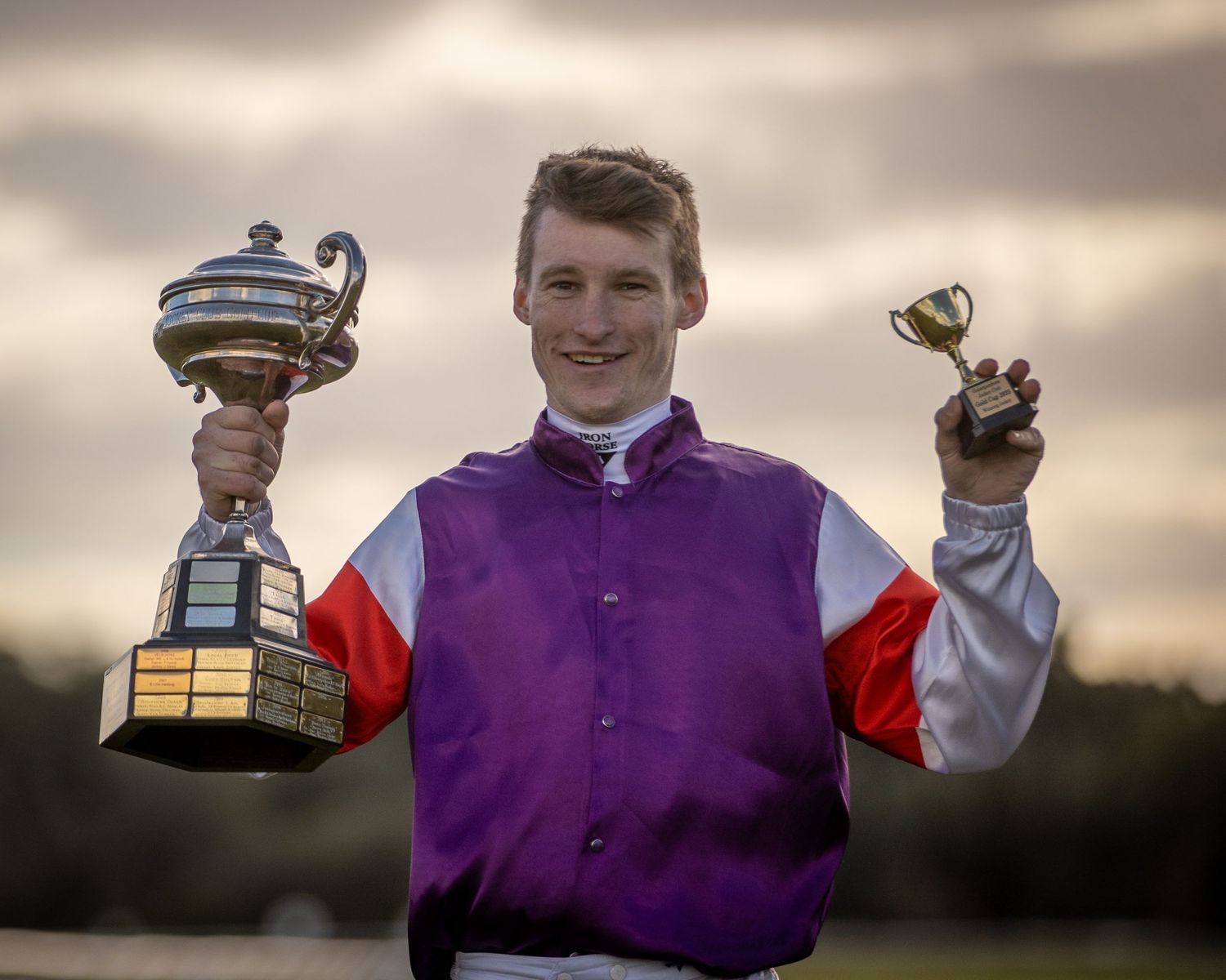 Jockey displaying racing trophies against a cloudy sunset sky.