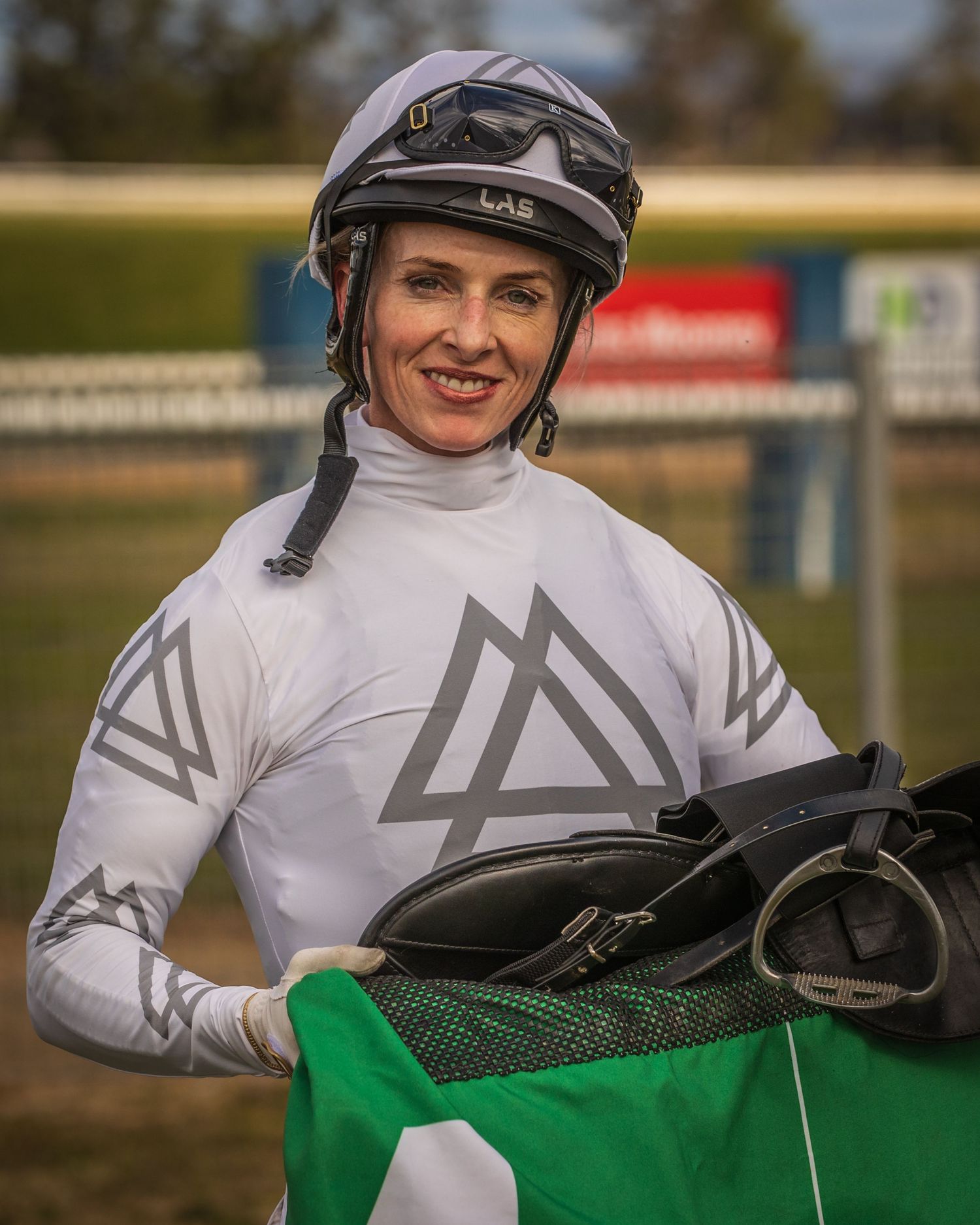 Smiling female jockey in white racing silks with geometric logo, holding a helmet at a racecourse.