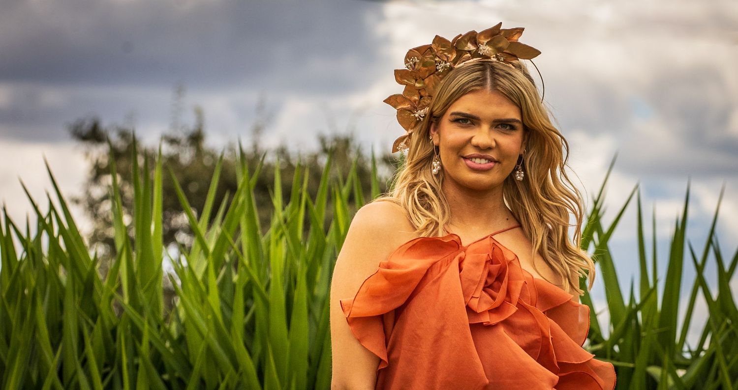 Woman in bright orange dress poses in tall green cornfield on cloudy summer day.