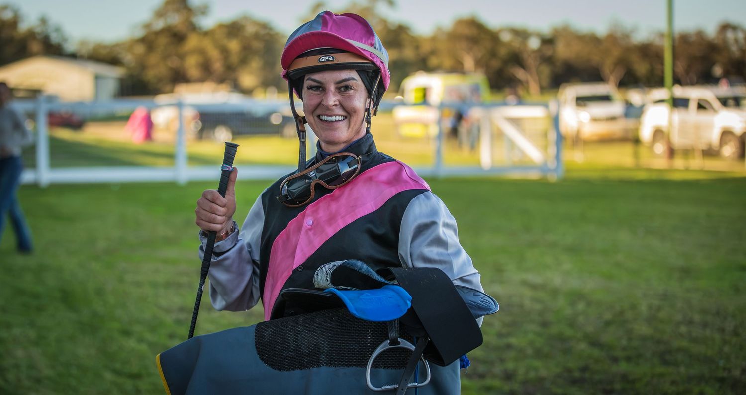 Smiling jockey in pink and grey silks holding a riding crop at a sunny racecourse.